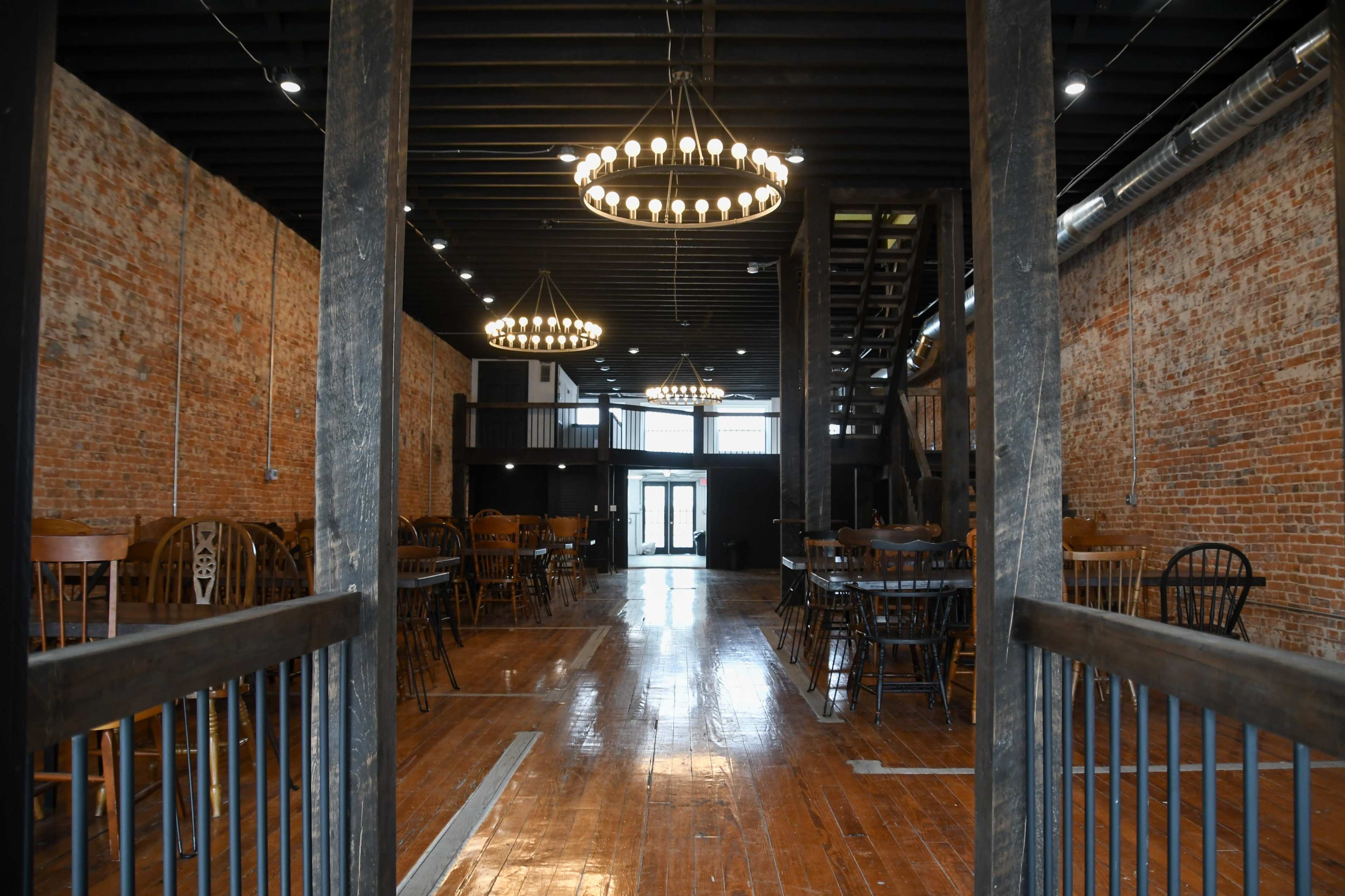 An interior view of a spacious venue featuring wooden flooring, brick walls, and multiple chandeliers hanging from the ceiling.