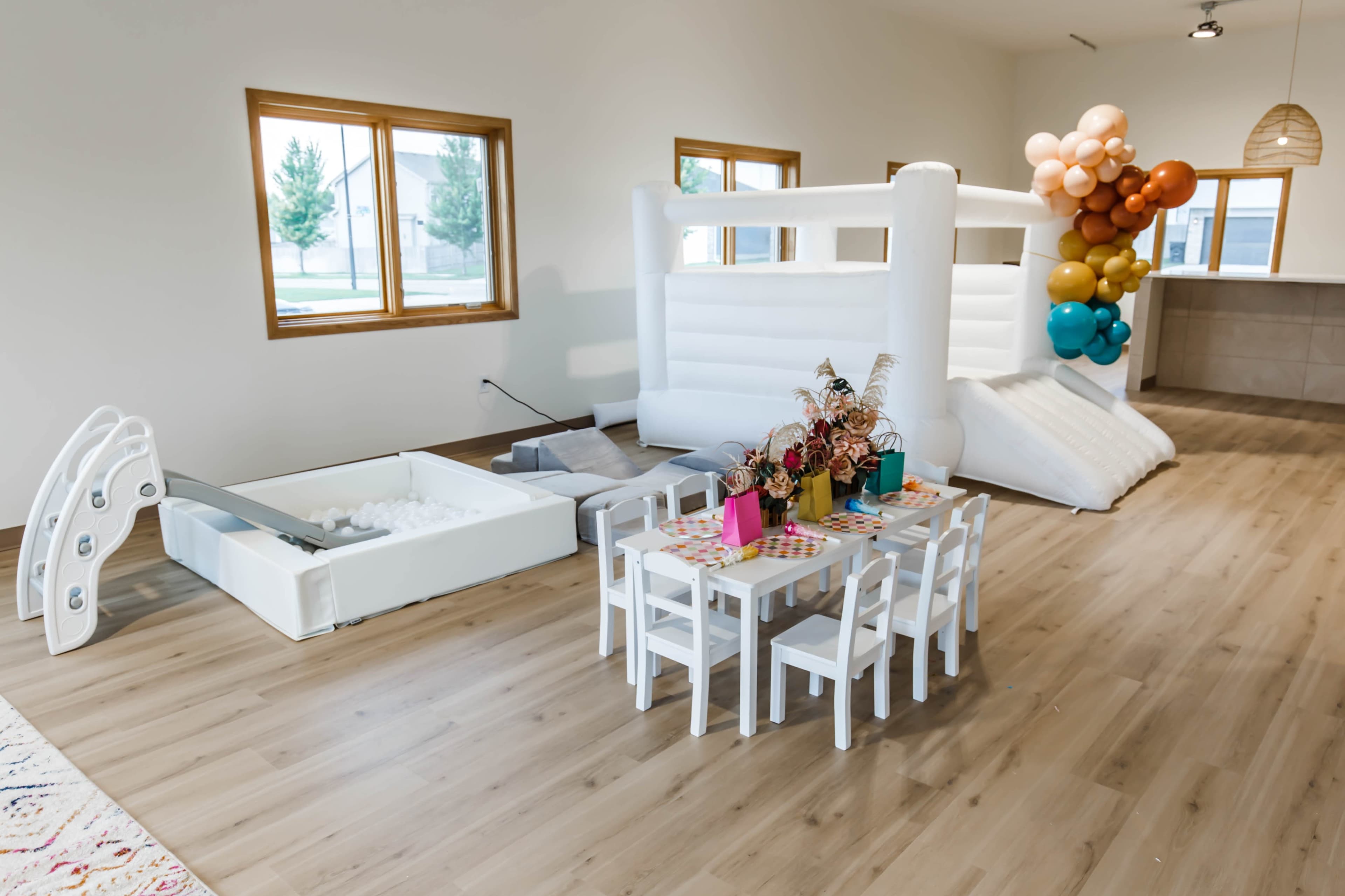 A playroom features a white inflatable slide and climbing structure alongside a small table with chairs set for children, surrounded by decorative balloons.