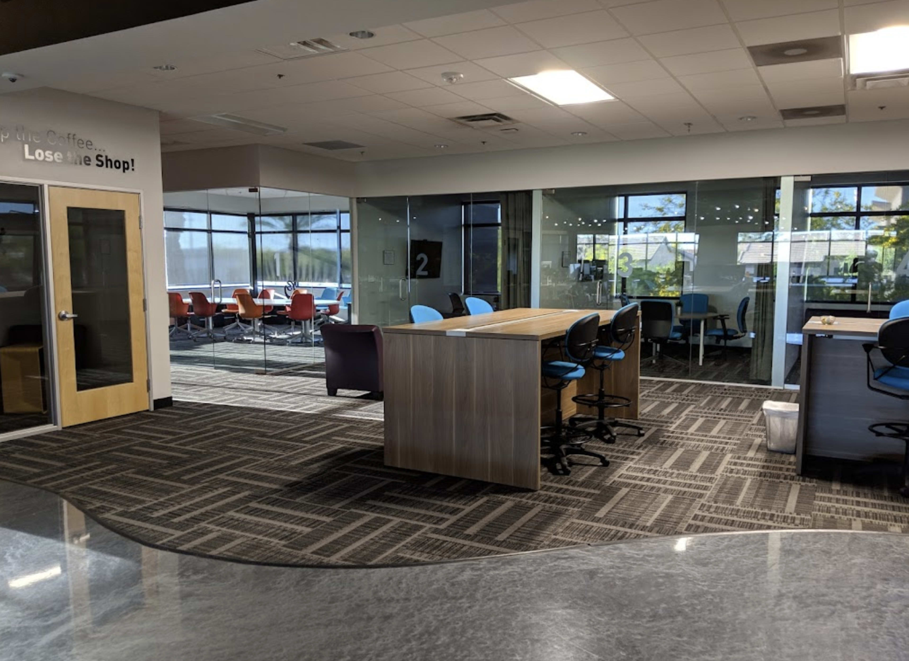 The image shows a modern office space with a communal table, chairs, and a lounge area, featuring glass walls that separate different sections.