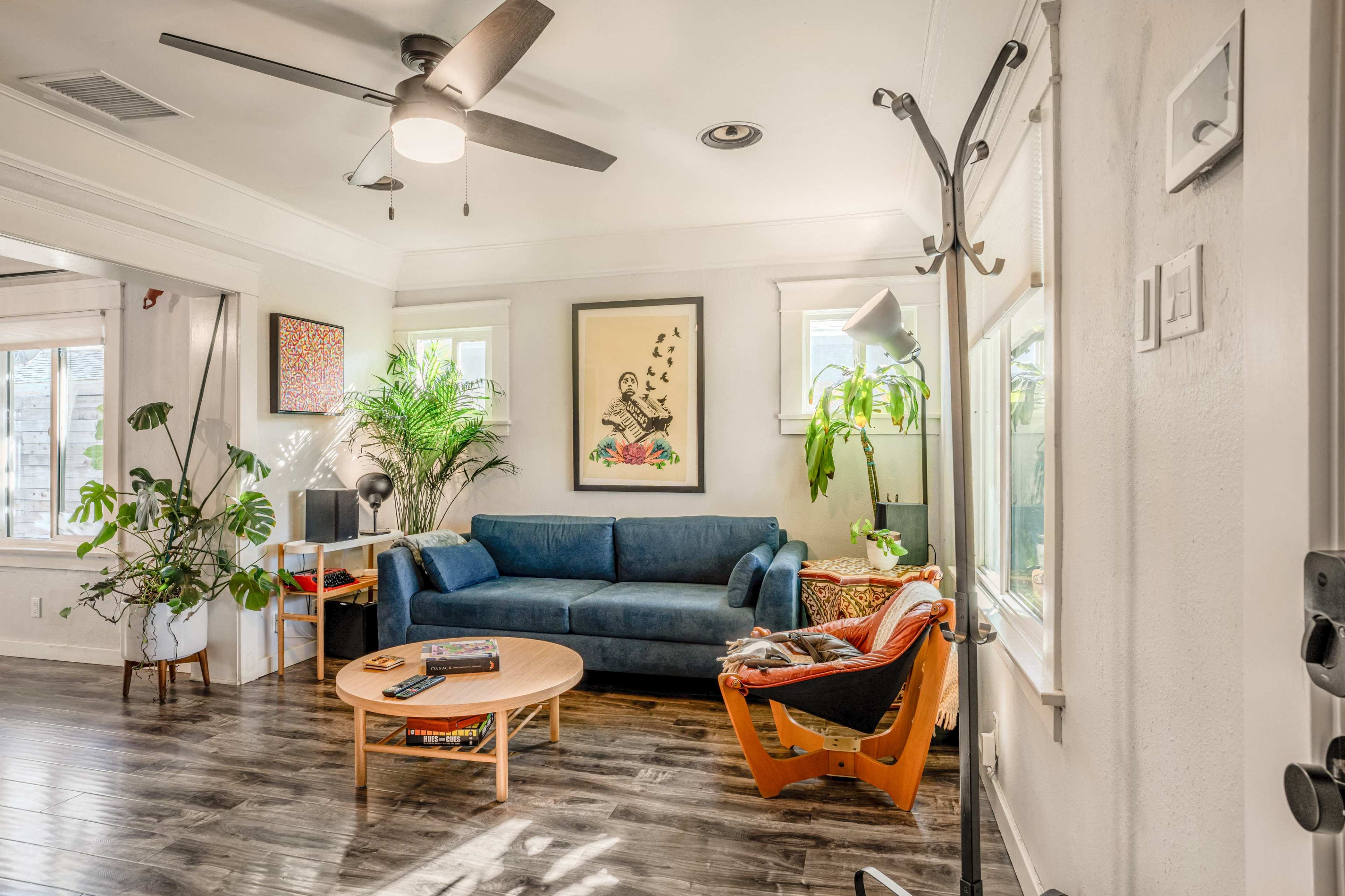 The image shows a living room with a blue sofa, a round wooden coffee table, plants, a wall-mounted coat rack, and natural light streaming through the windows.