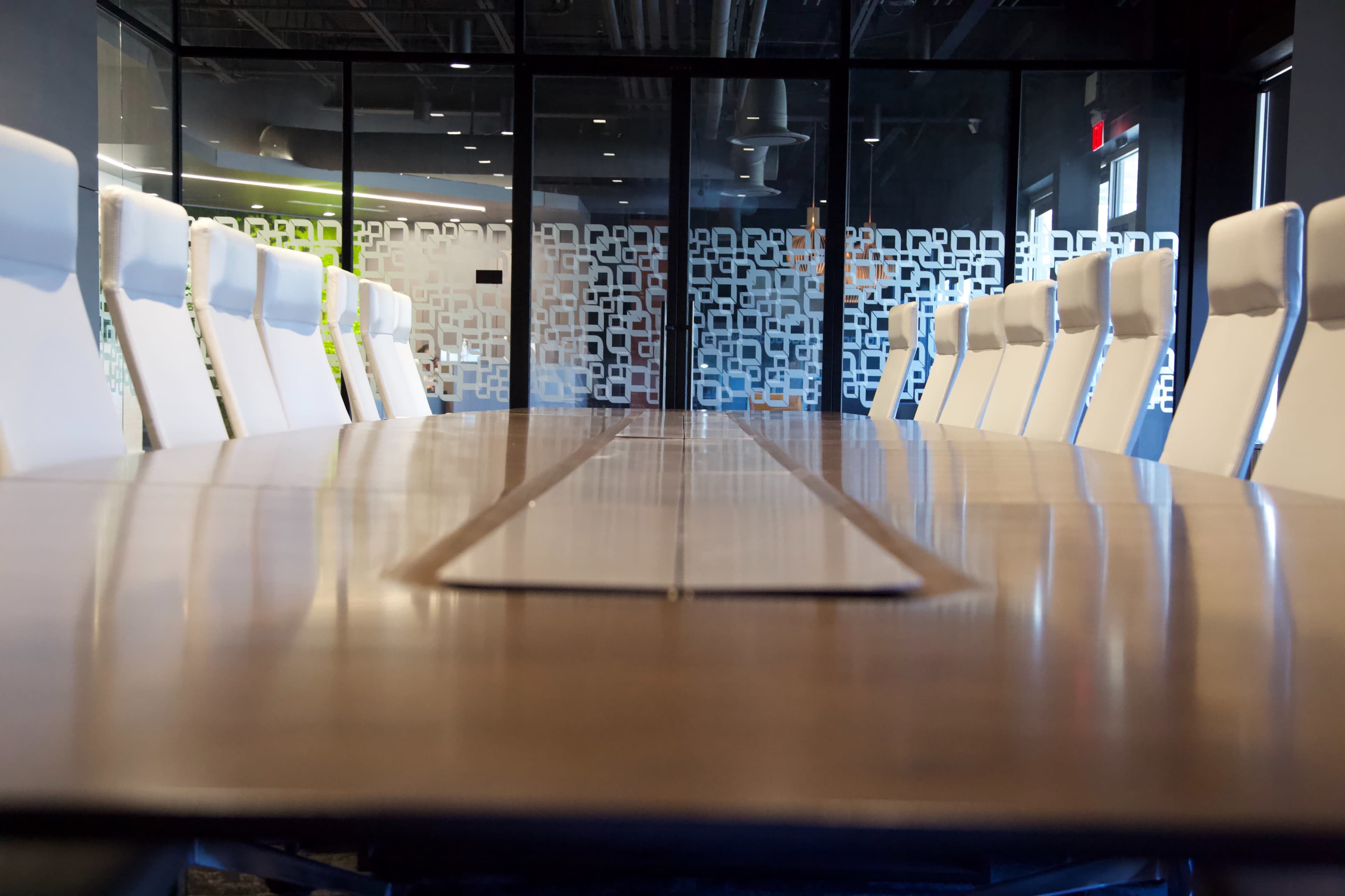 A long wooden conference table is lined with white chairs in a modern boardroom, with glass walls displaying a geometric pattern.