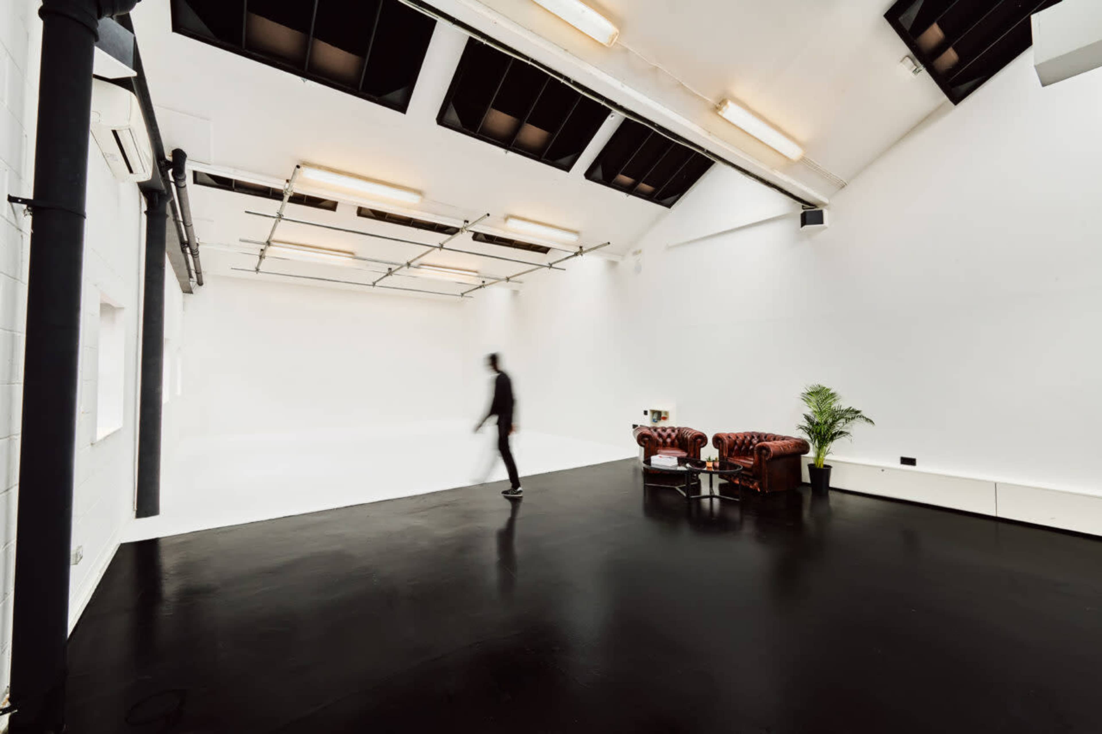 A person walks through a spacious, minimalist studio featuring a black floor and a brown leather couch.