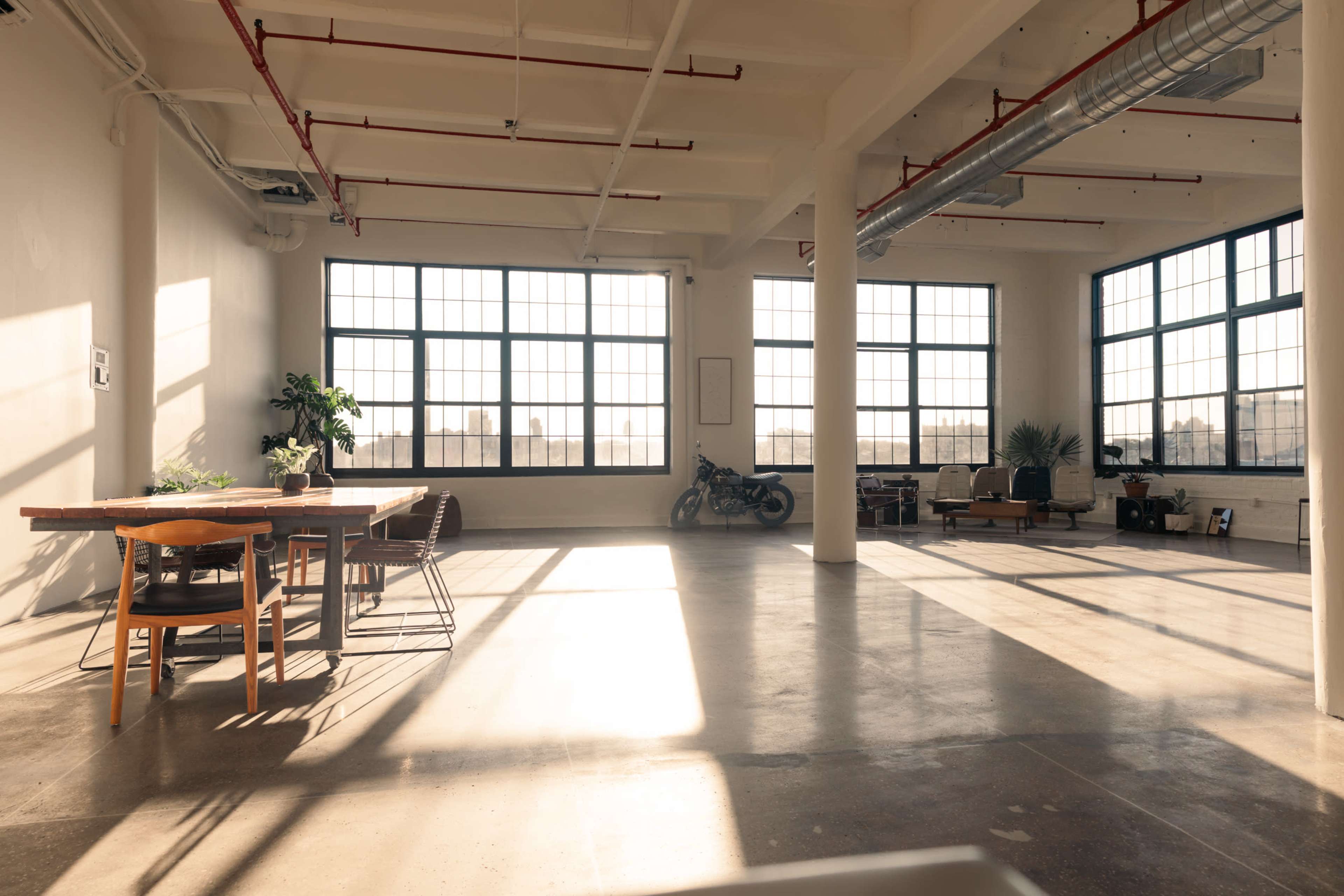 The image shows a spacious, sunlit loft with large windows, a dining table, chairs, and a motorcycle in the corner.