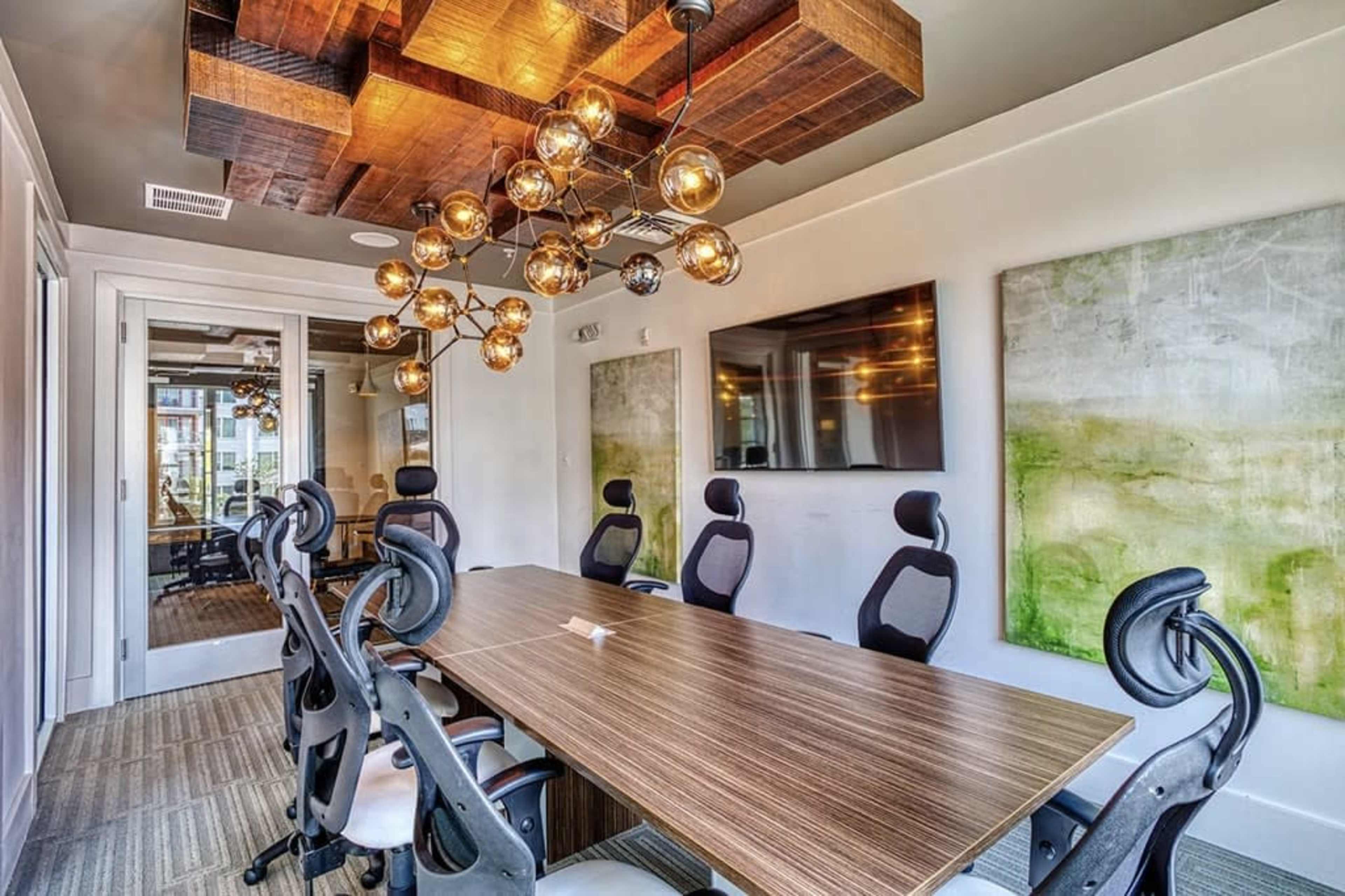 The image shows a modern conference room with a long wooden table, ergonomic chairs, artistic wall decor, and unique overhead lighting.