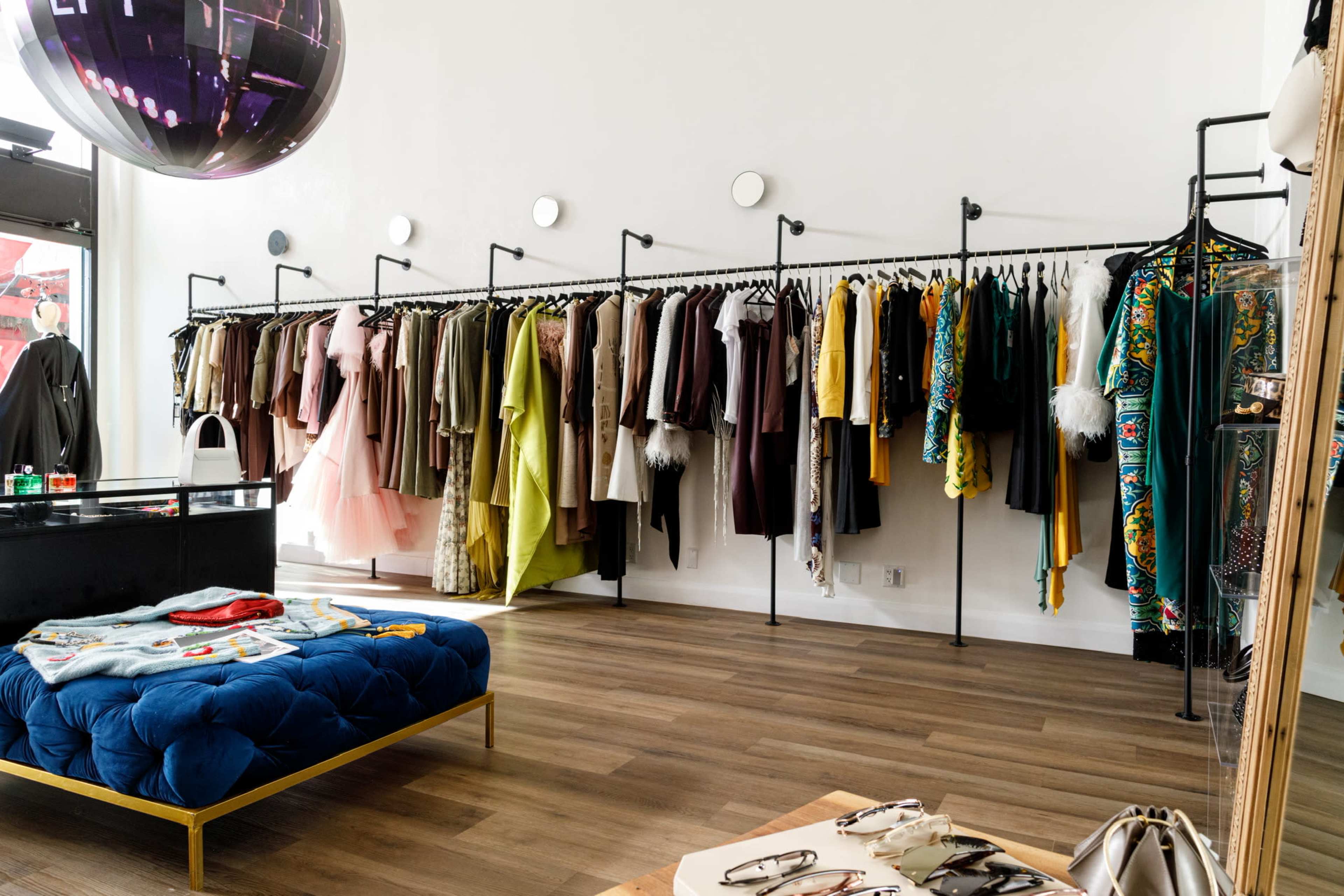 The image shows a boutique interior with a display of colorful clothing hanging on racks along the wall and a blue upholstered bench in the center.