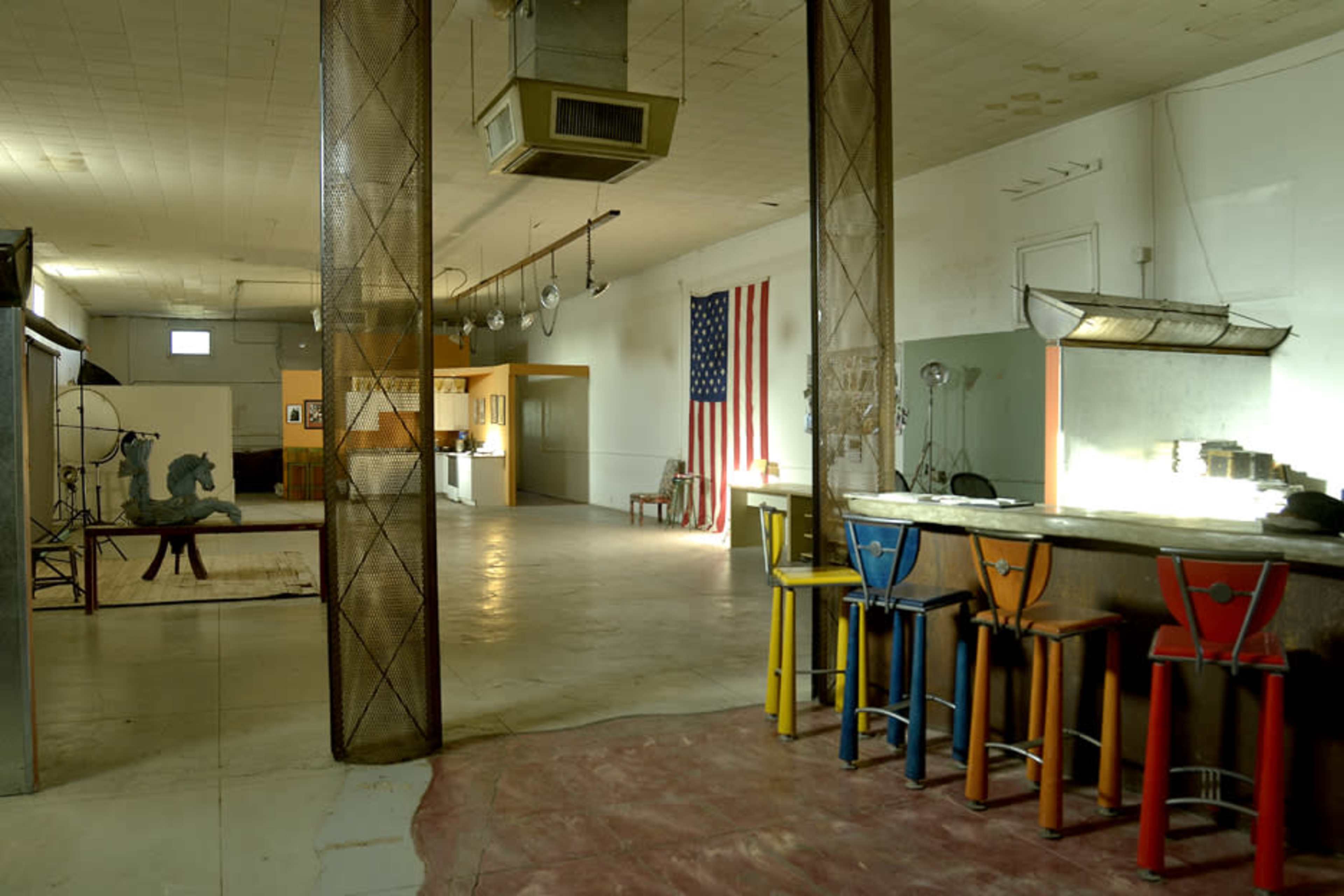 A spacious, partially empty interior featuring a bar with colorful stools, an American flag hanging on the wall, and various objects scattered throughout the room.