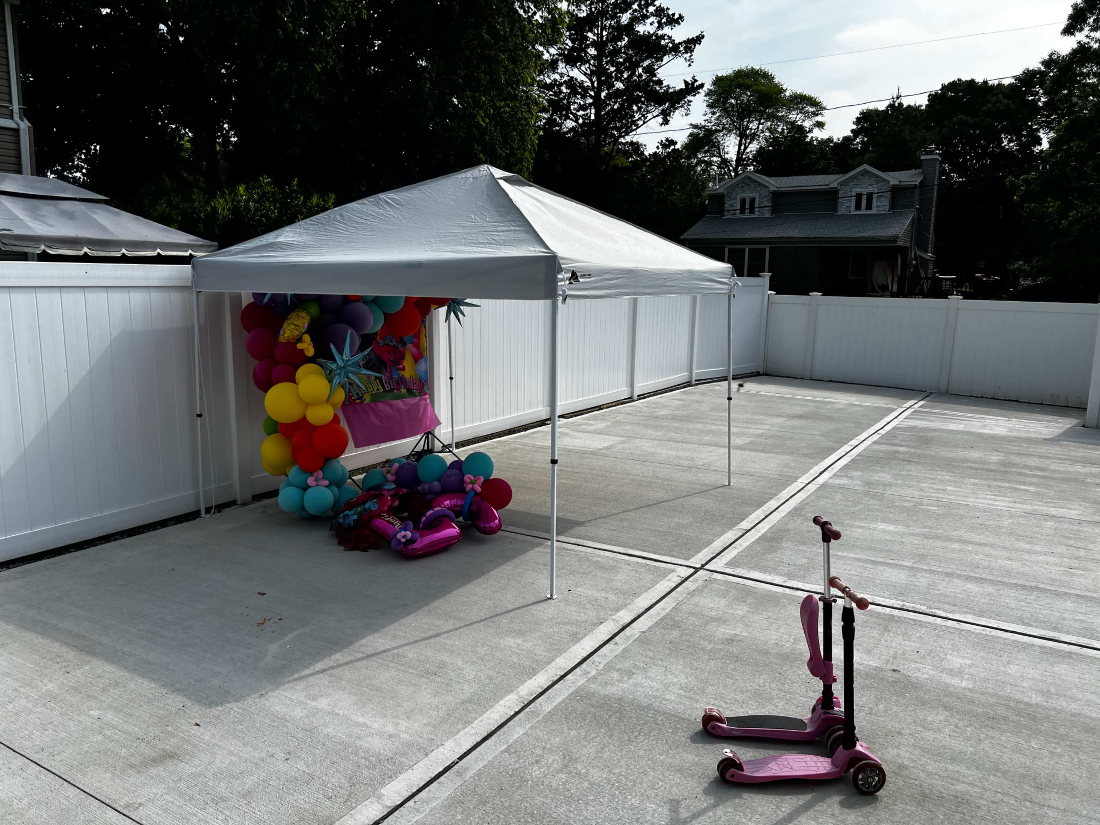 A white canopy tent with colorful balloons is set up in an empty concrete area beside two pink scooters.