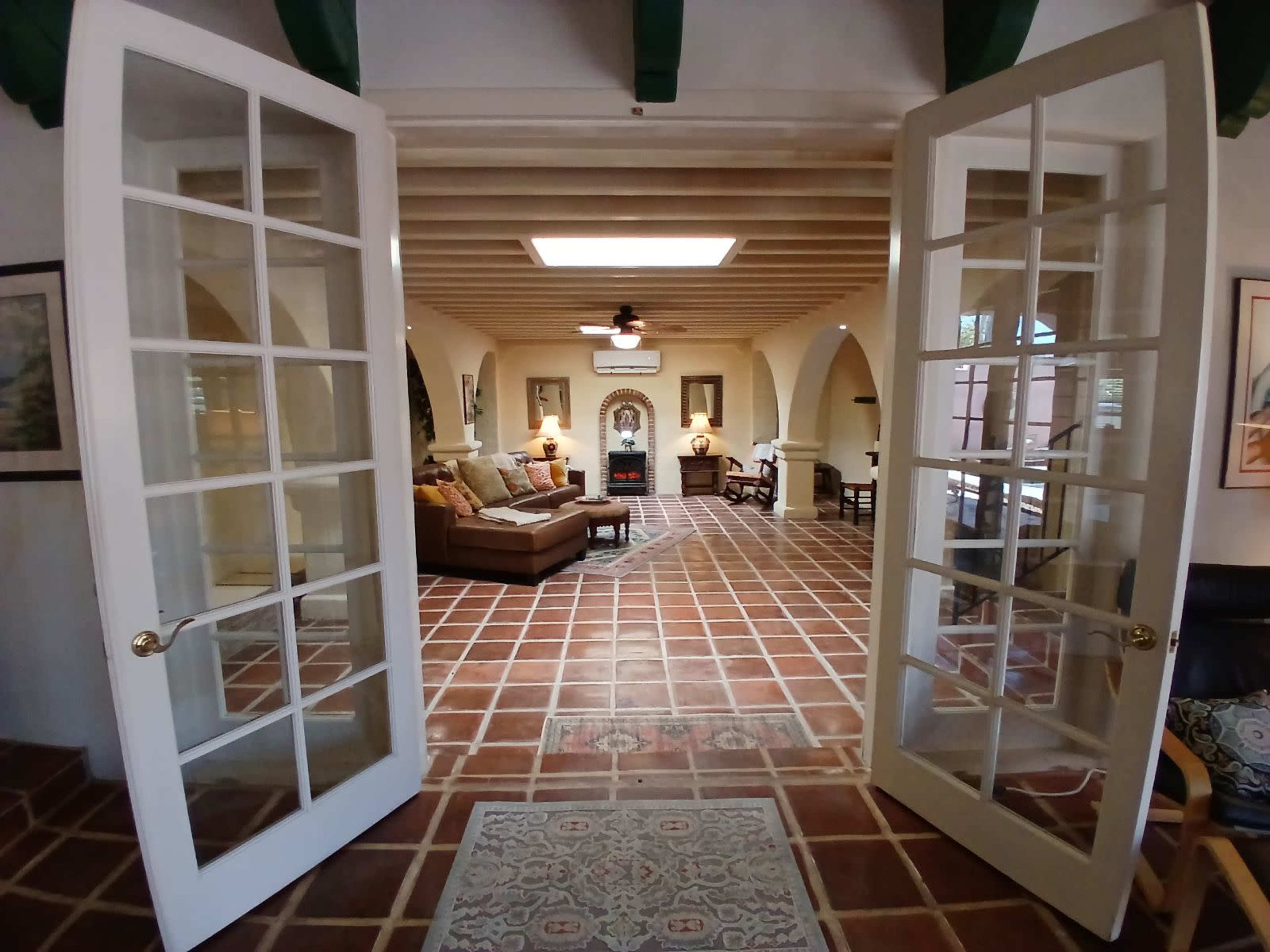 A spacious living room viewed through double French doors, featuring tiled floors, a central area rug, and furniture arranged around a fireplace.