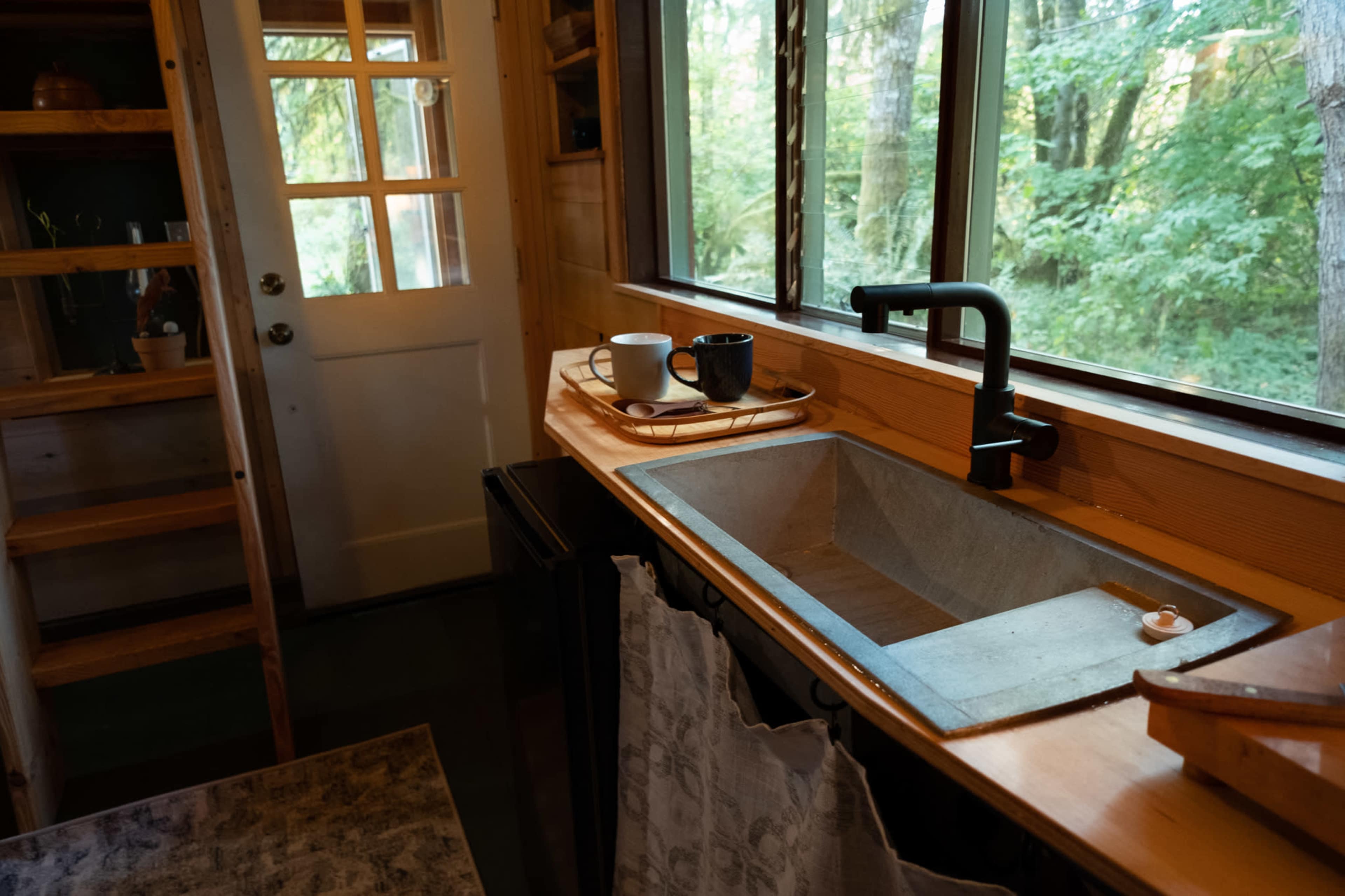 A small kitchen area with a concrete sink, wooden countertop, and large windows overlooking a green landscape.