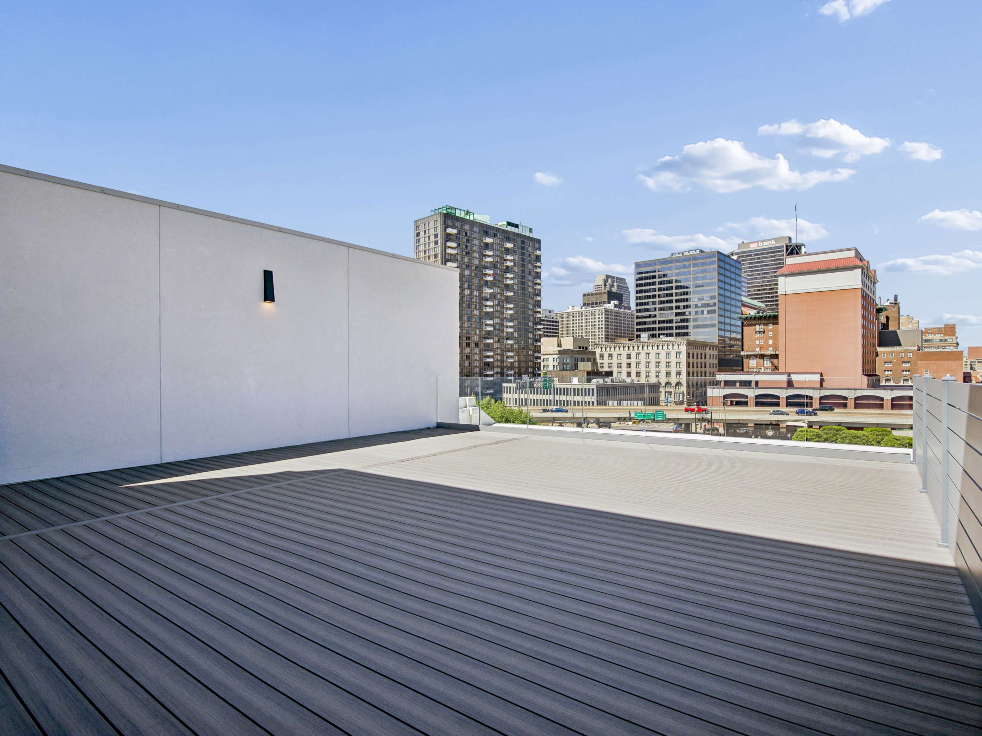 The image shows a spacious rooftop terrace with a wooden deck, overlooking a city skyline featuring various high-rise buildings under a clear blue sky.