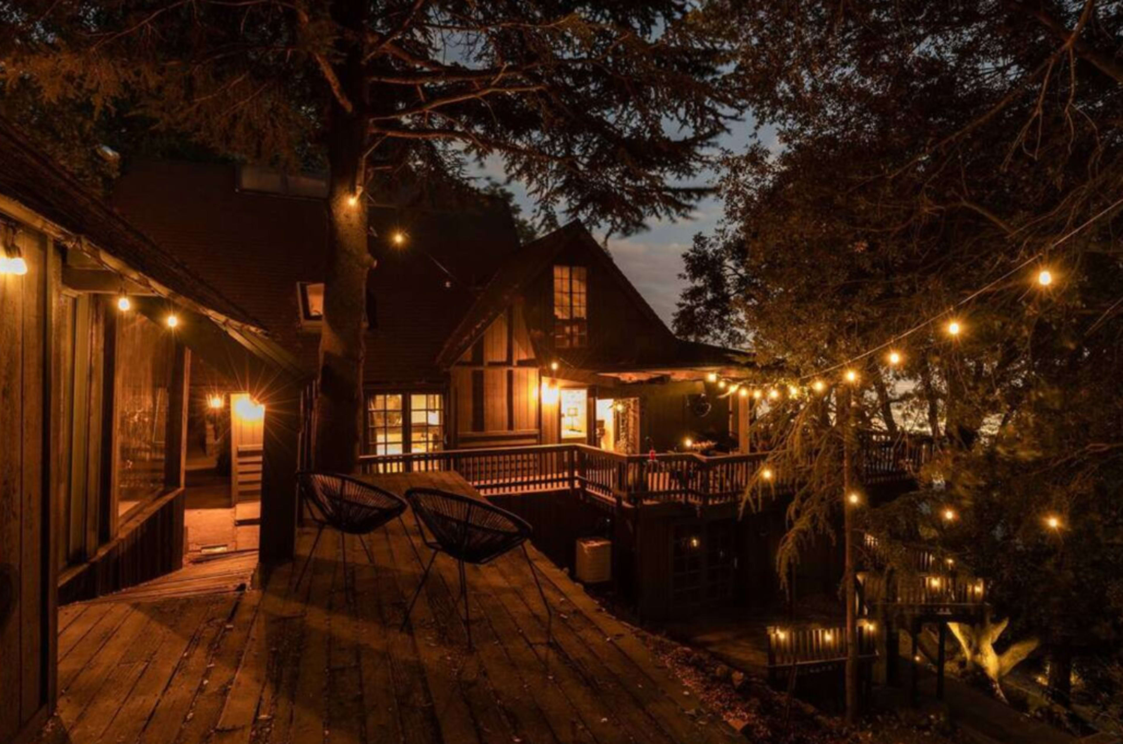 A wooden deck with two chairs overlooks a house illuminated by string lights at dusk, surrounded by trees.