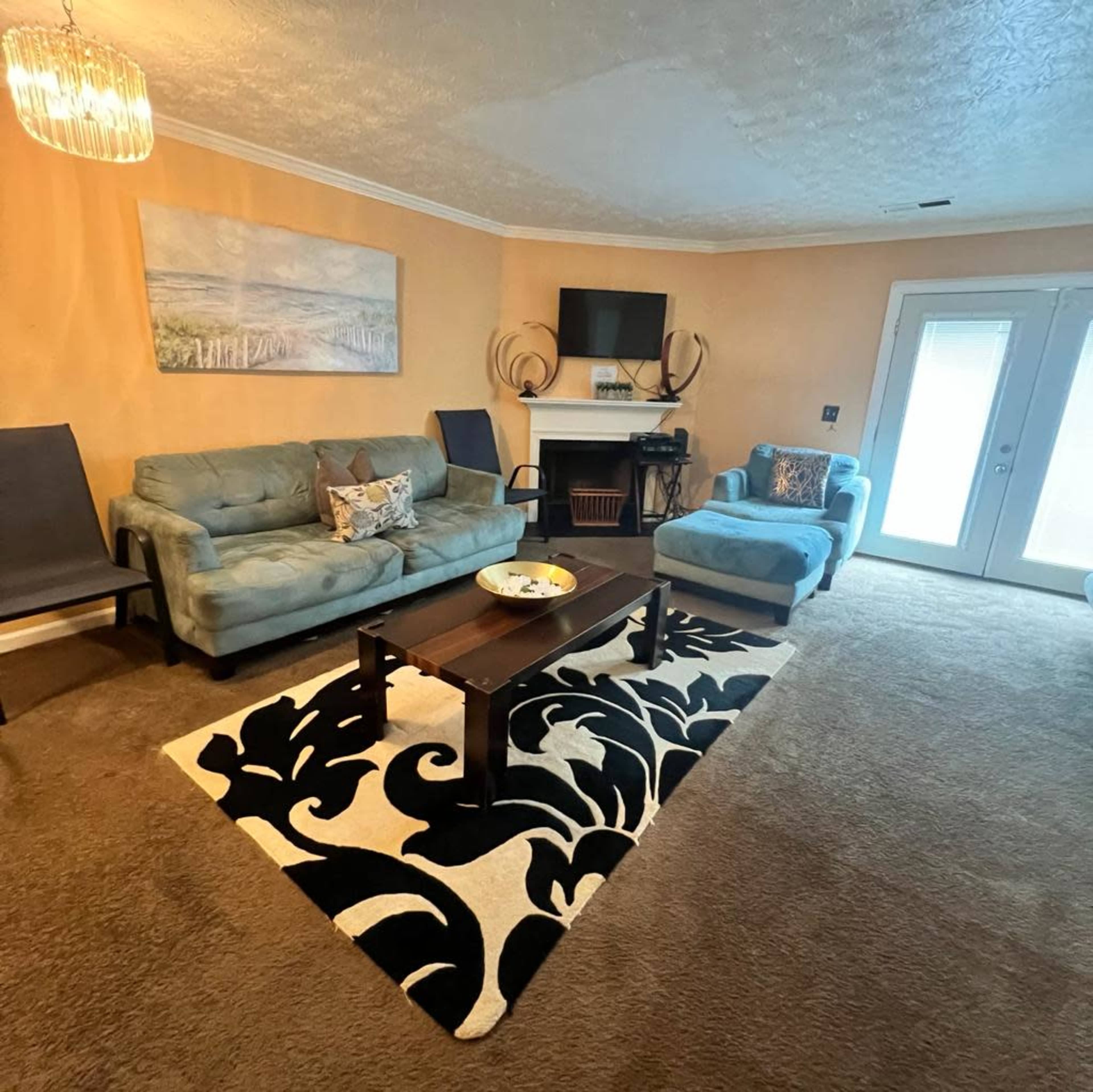 The living room features two sofas, a coffee table, a TV mounted above a fireplace, and a large area rug on carpet flooring.