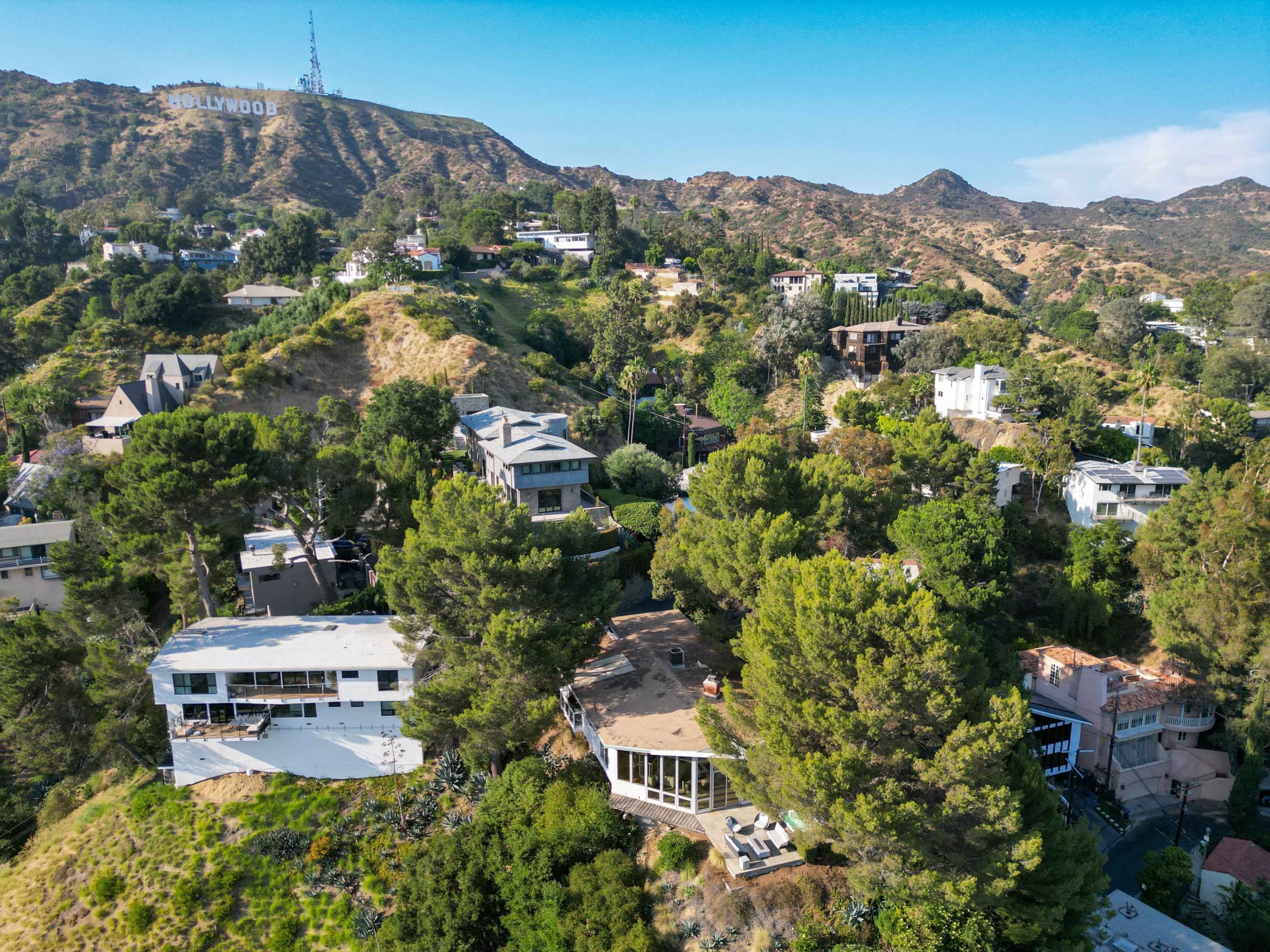 The image shows a hillside neighborhood in Los Angeles with houses surrounded by greenery and the Hollywood sign visible in the background.