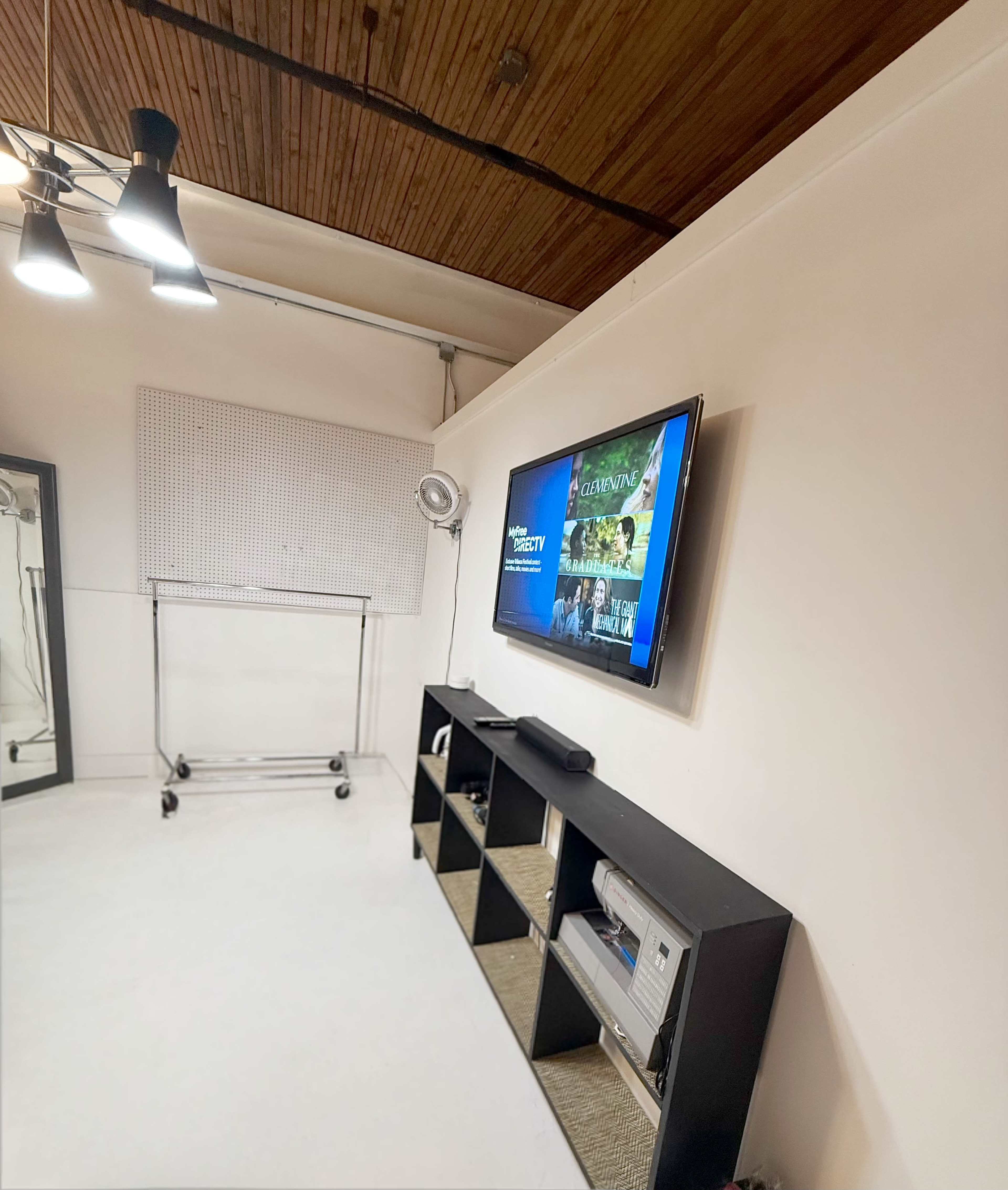 A flat-screen television is mounted on a white wall beside a black shelving unit in a brightly lit room with wooden ceiling panels.