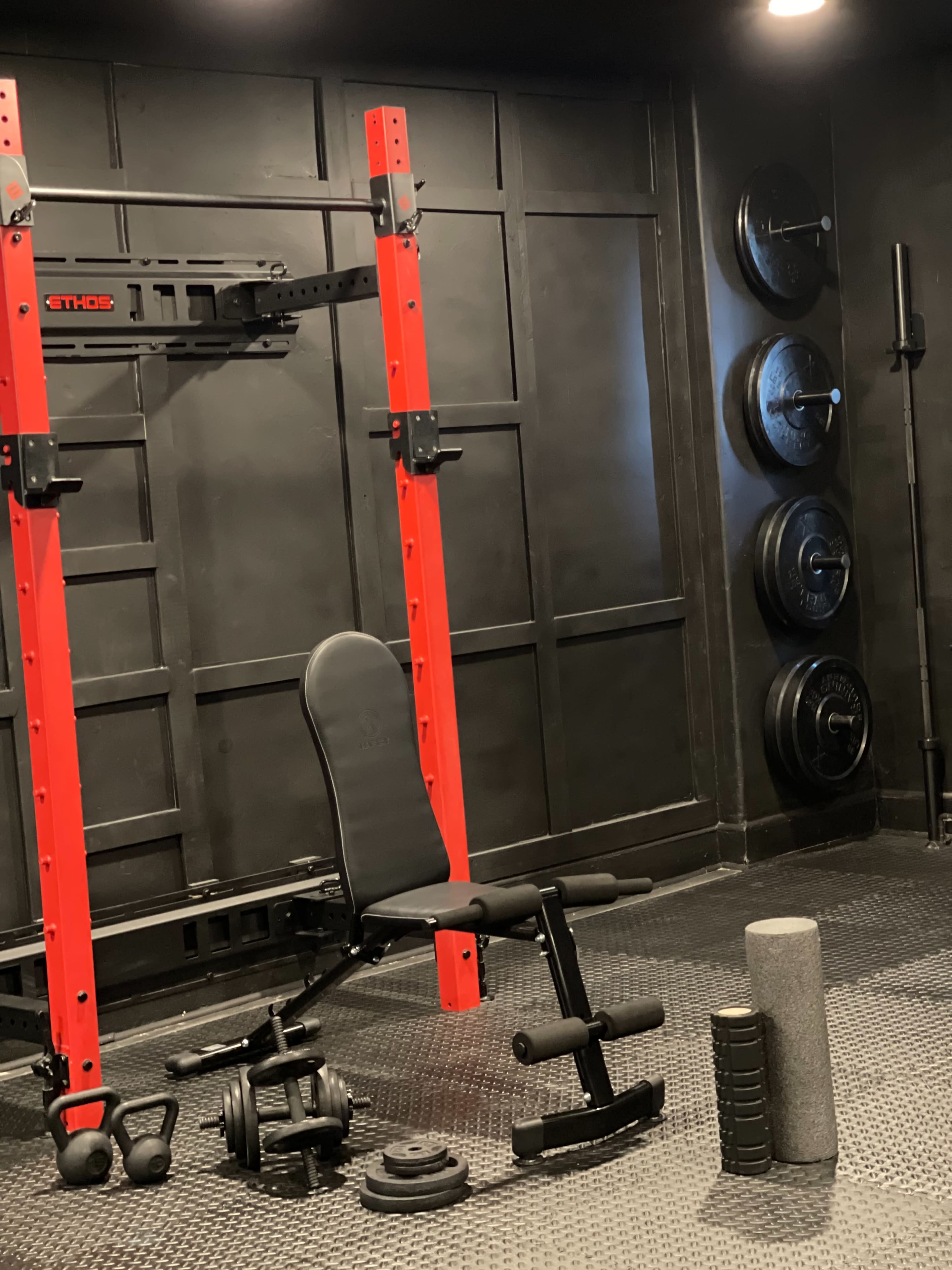 A black-walled gym area featuring a red power rack, adjustable workout bench, dumbbells, weight plates, and a foam roller arranged on a rubber floor mat.