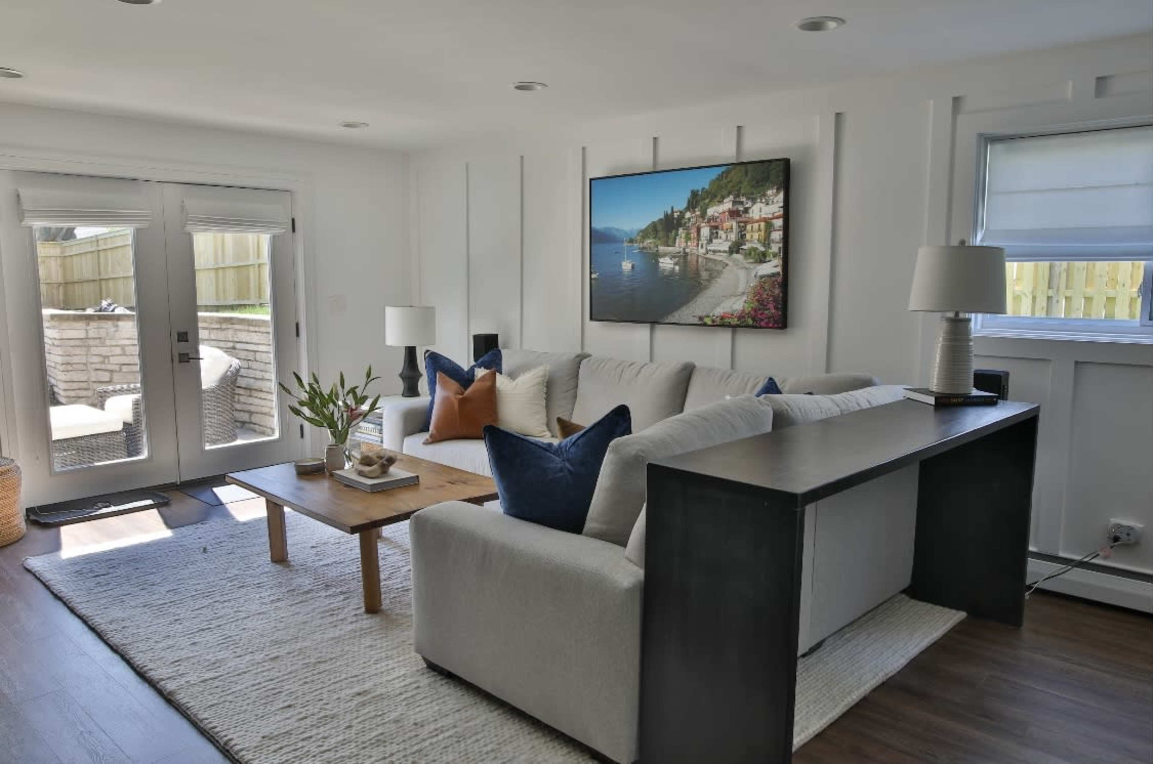 A modern living room with a white sofa, a wooden coffee table, and a large wall-mounted painting, featuring a view of a coastal village.