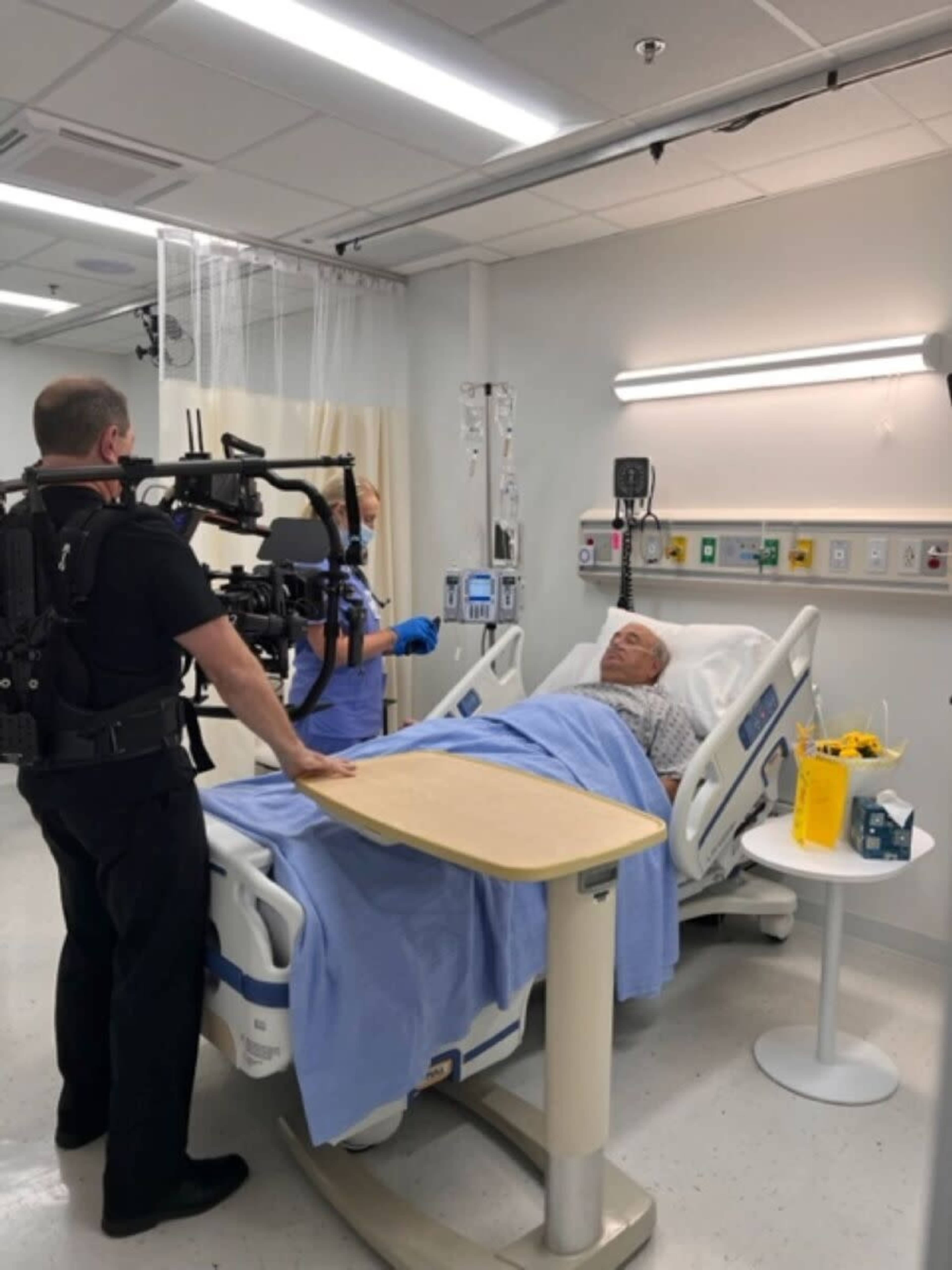 A patient lies on a hospital bed while a medical team uses a camera rig to film the scene in a healthcare setting.