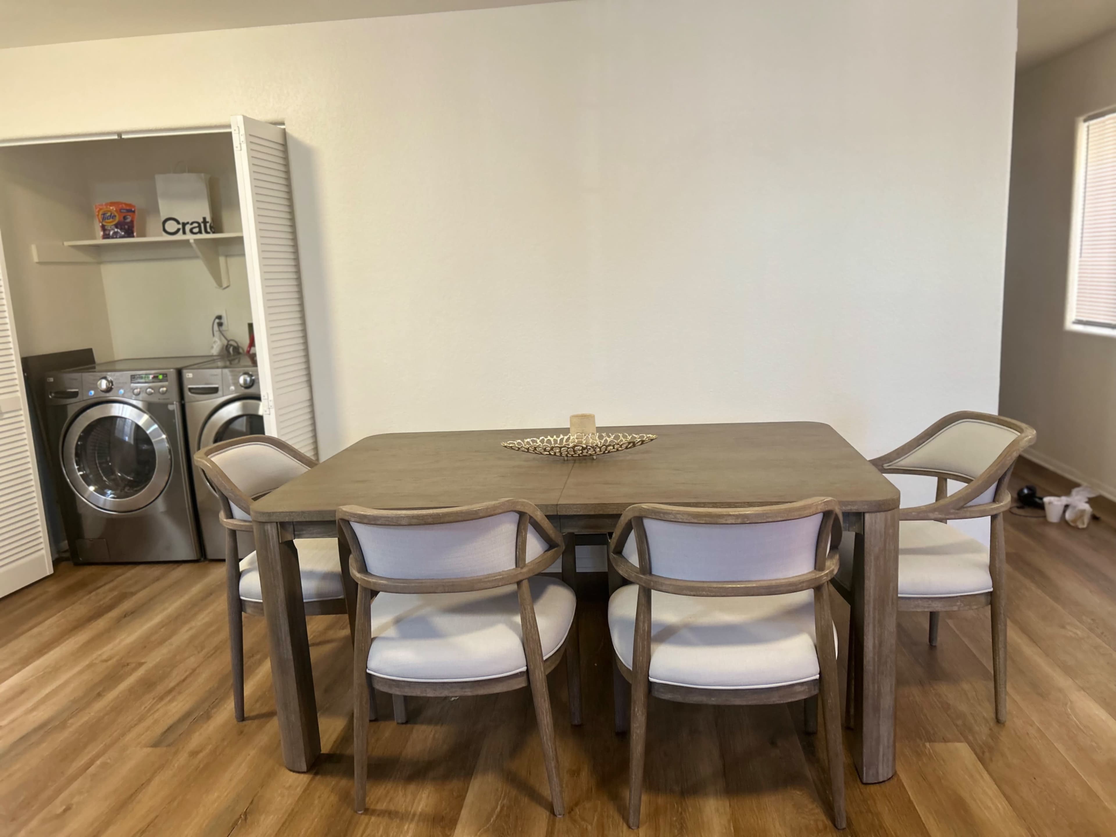 A wooden dining table with four chairs is set in a room featuring a small laundry area in the background.