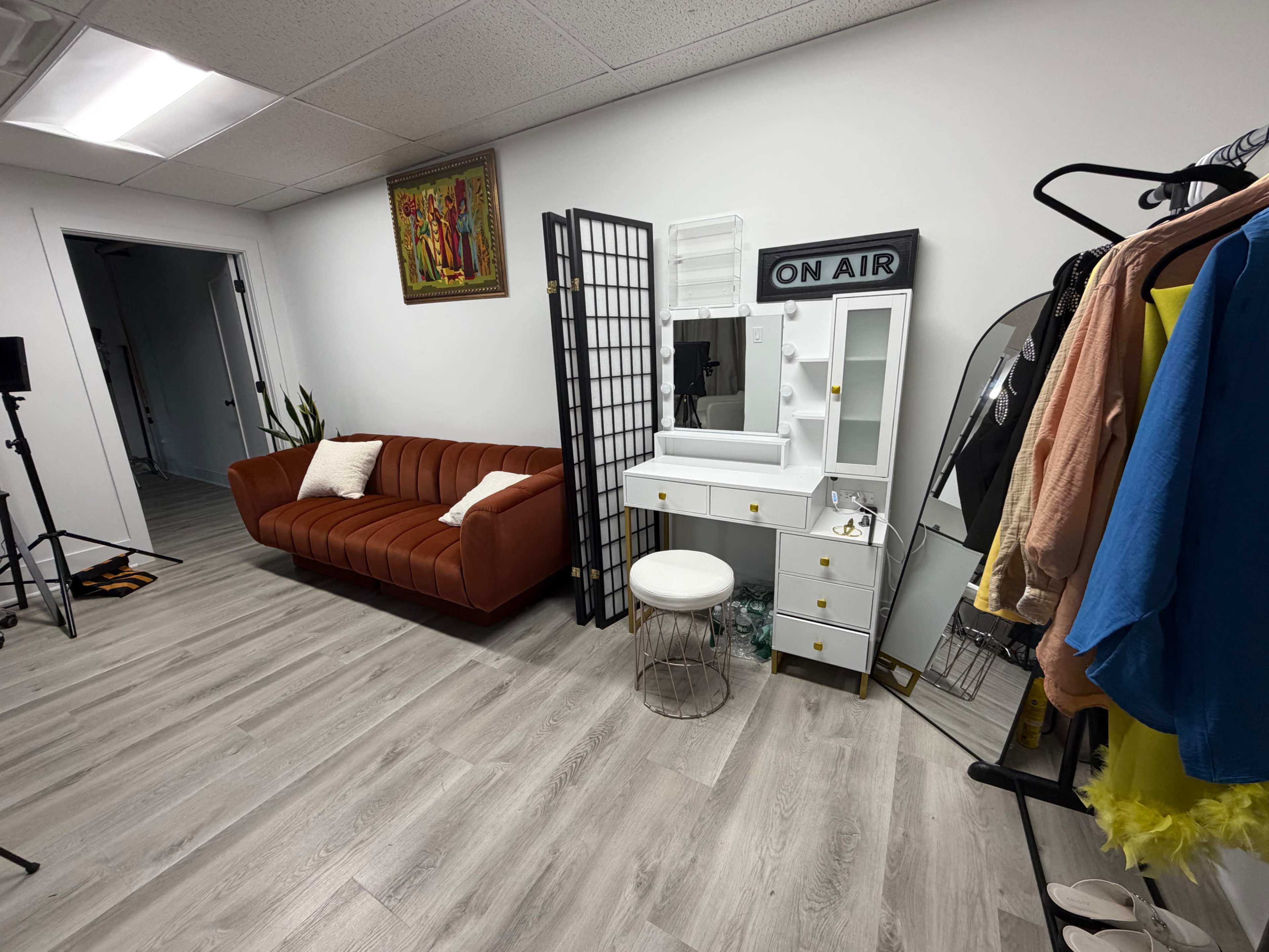 The image shows a modern studio setup featuring a brown sofa, a makeup vanity with a mirror, a folding screen, and various clothing items hanging on a rack.