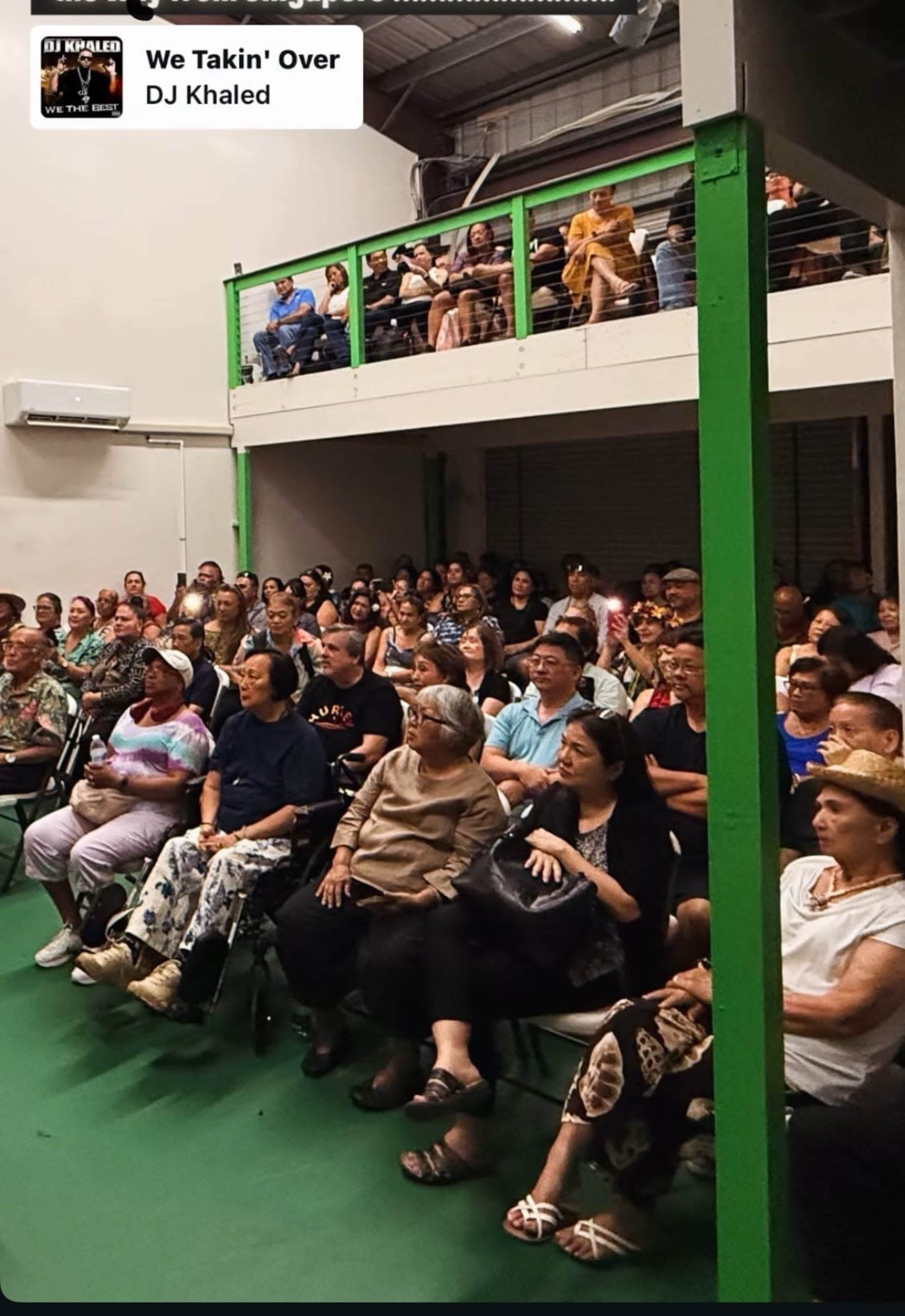 A large audience consisting of men and women sits in a spacious venue, with some people in an upper seating area observing the event below.