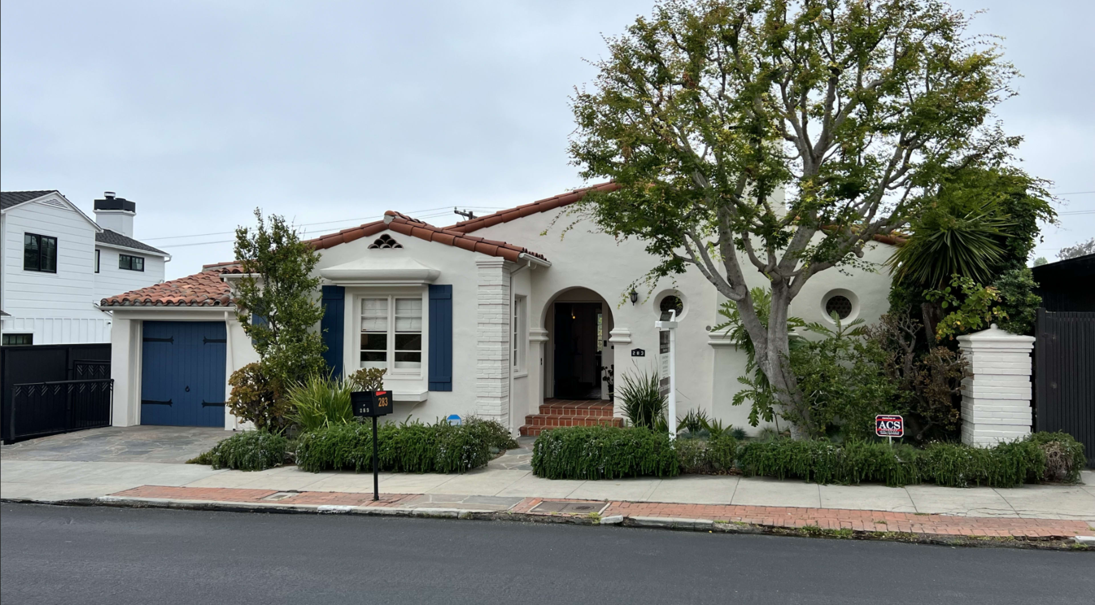 A single-story, stucco house with a red-tiled roof features blue shutters and a lush garden in front.