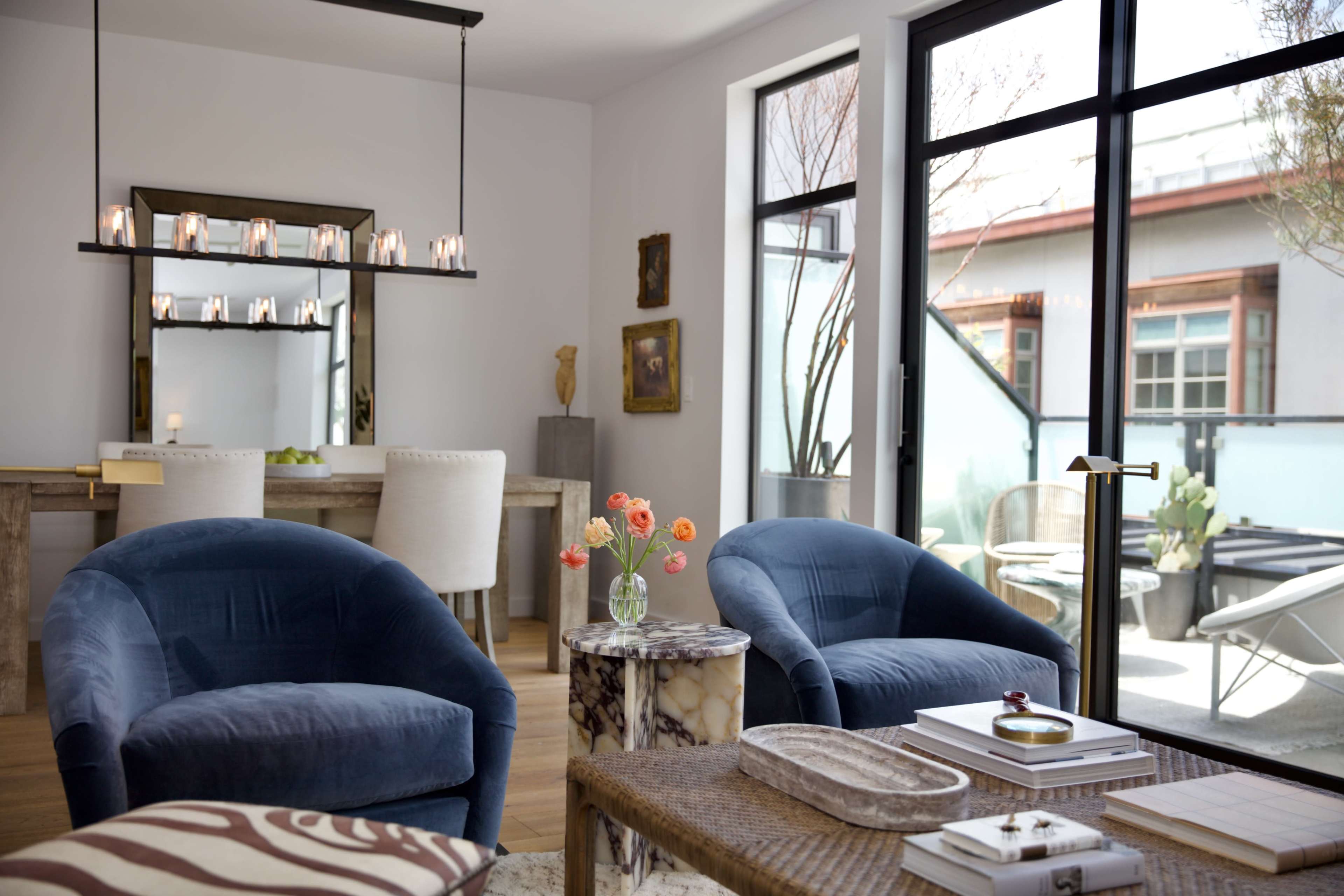 A modern living room with two blue armchairs facing a small round table, a dining area in the background, and large windows letting in natural light.