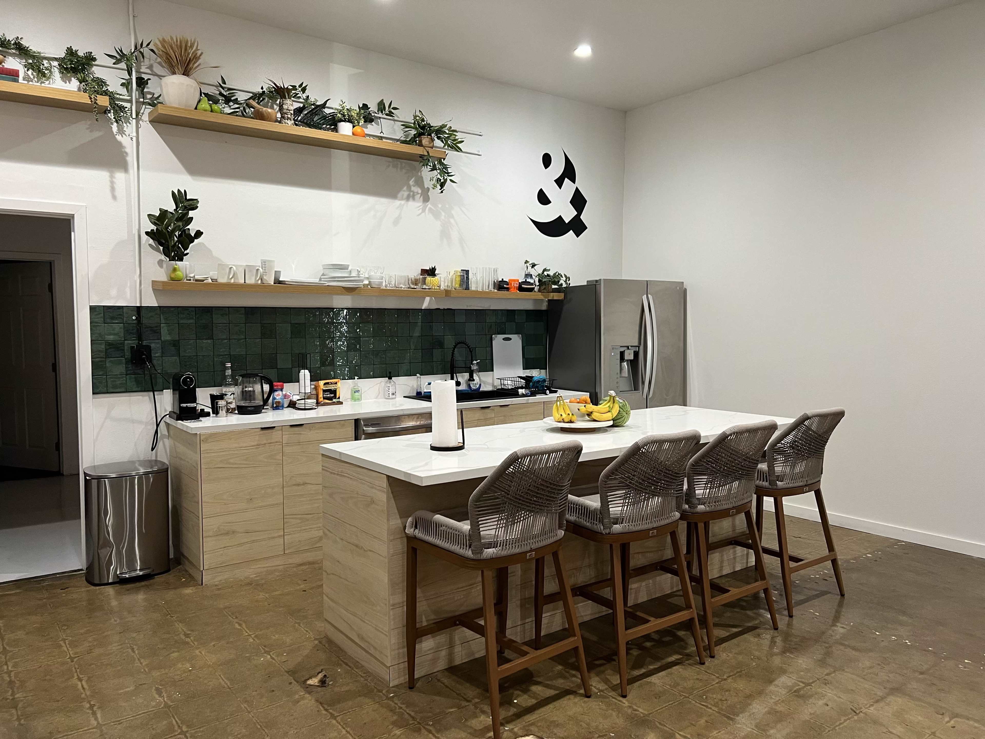 The kitchen features a central island with four chairs, green tiled backsplash, and open shelves holding various kitchen items.