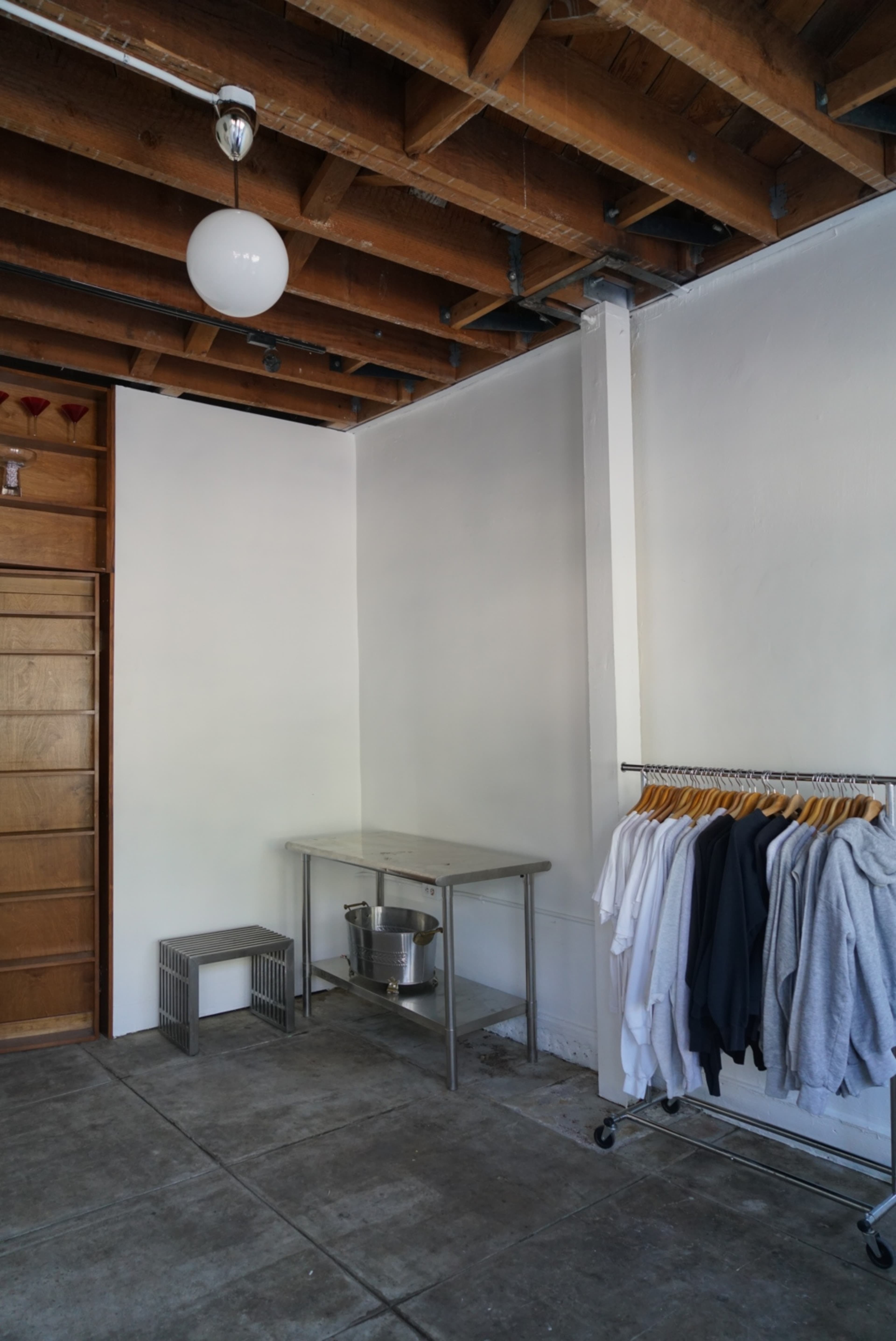 A minimalist interior space with exposed wooden beams, a metal table, a clothing rack displaying sweatshirts, and a concrete floor.