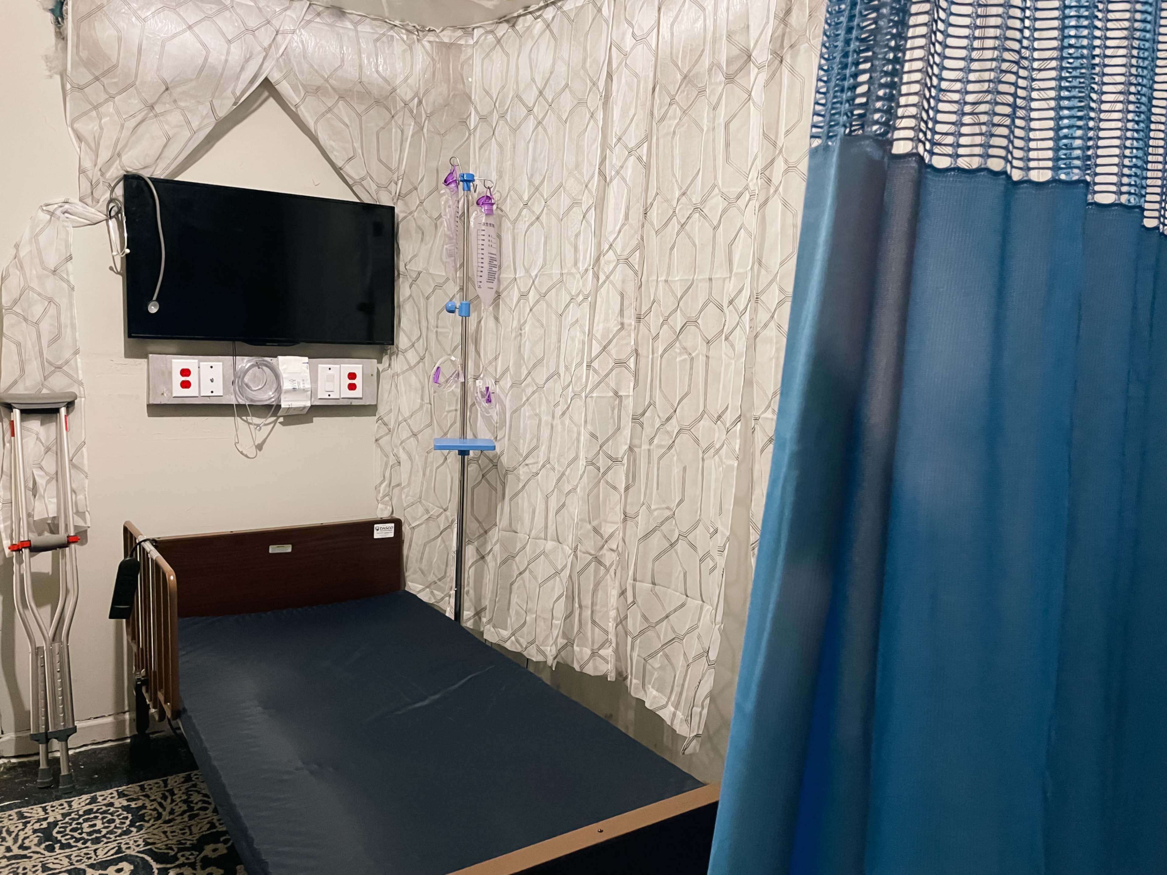 The image shows a hospital room with a bed, a television mounted on the wall, an IV stand, and curtains dividing the space.