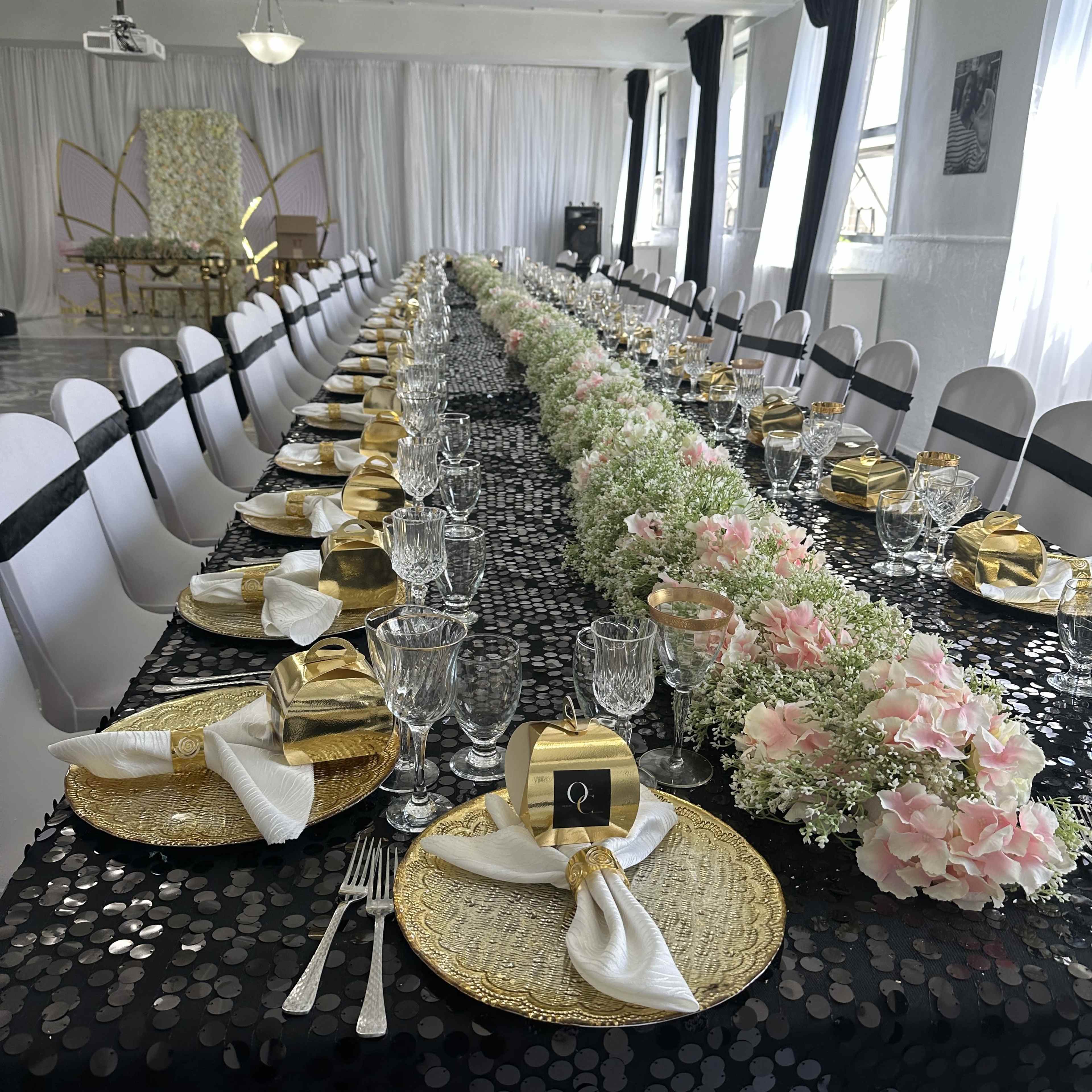 A long banquet table is elegantly set with gold-wrapped favors, crystal glasses, and floral centerpieces under a bright, airy venue.