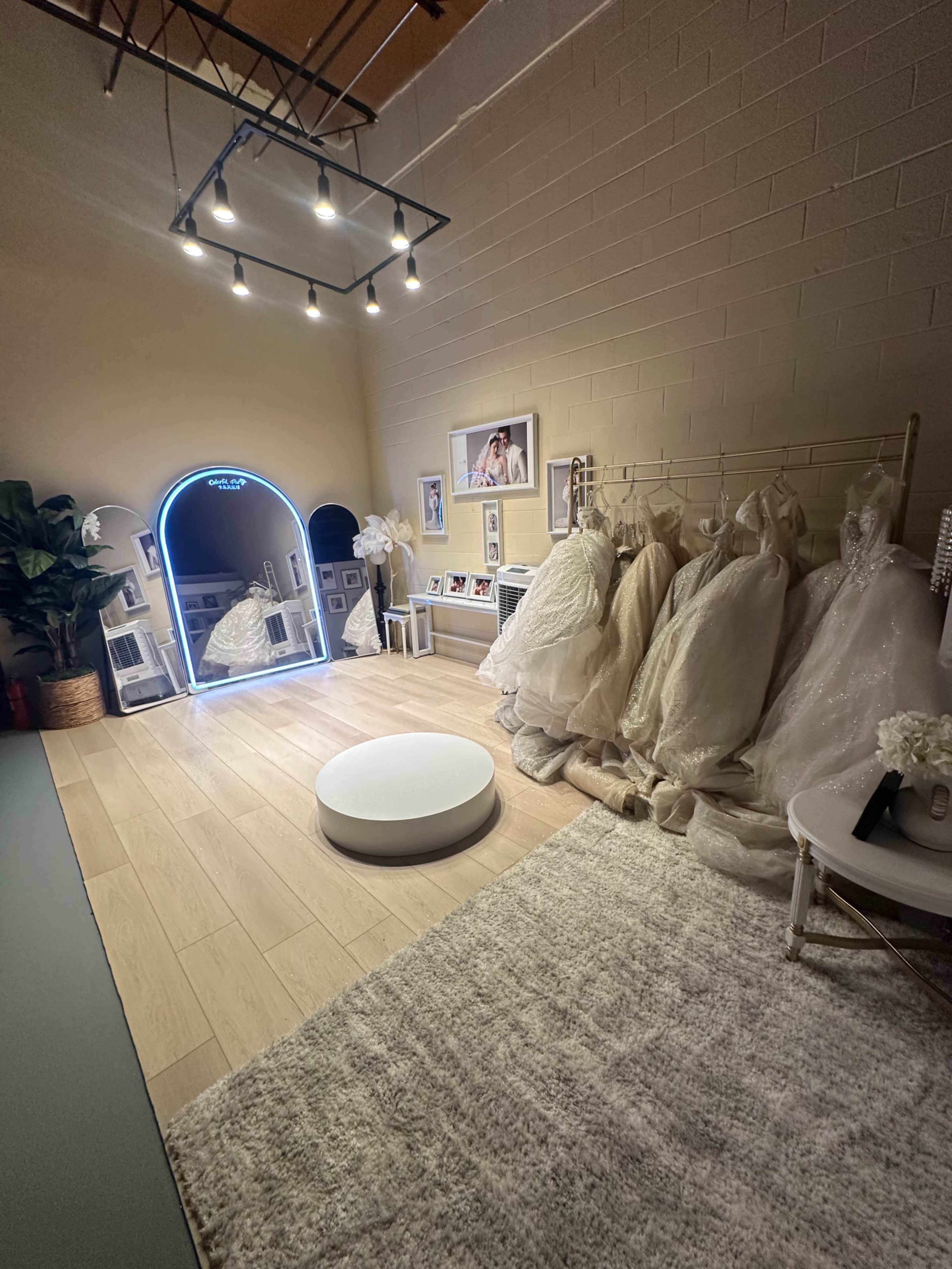 The image shows a well-lit boutique room featuring a circular display platform, a neon mirror, racks of wedding dresses, and elegant decor elements.
