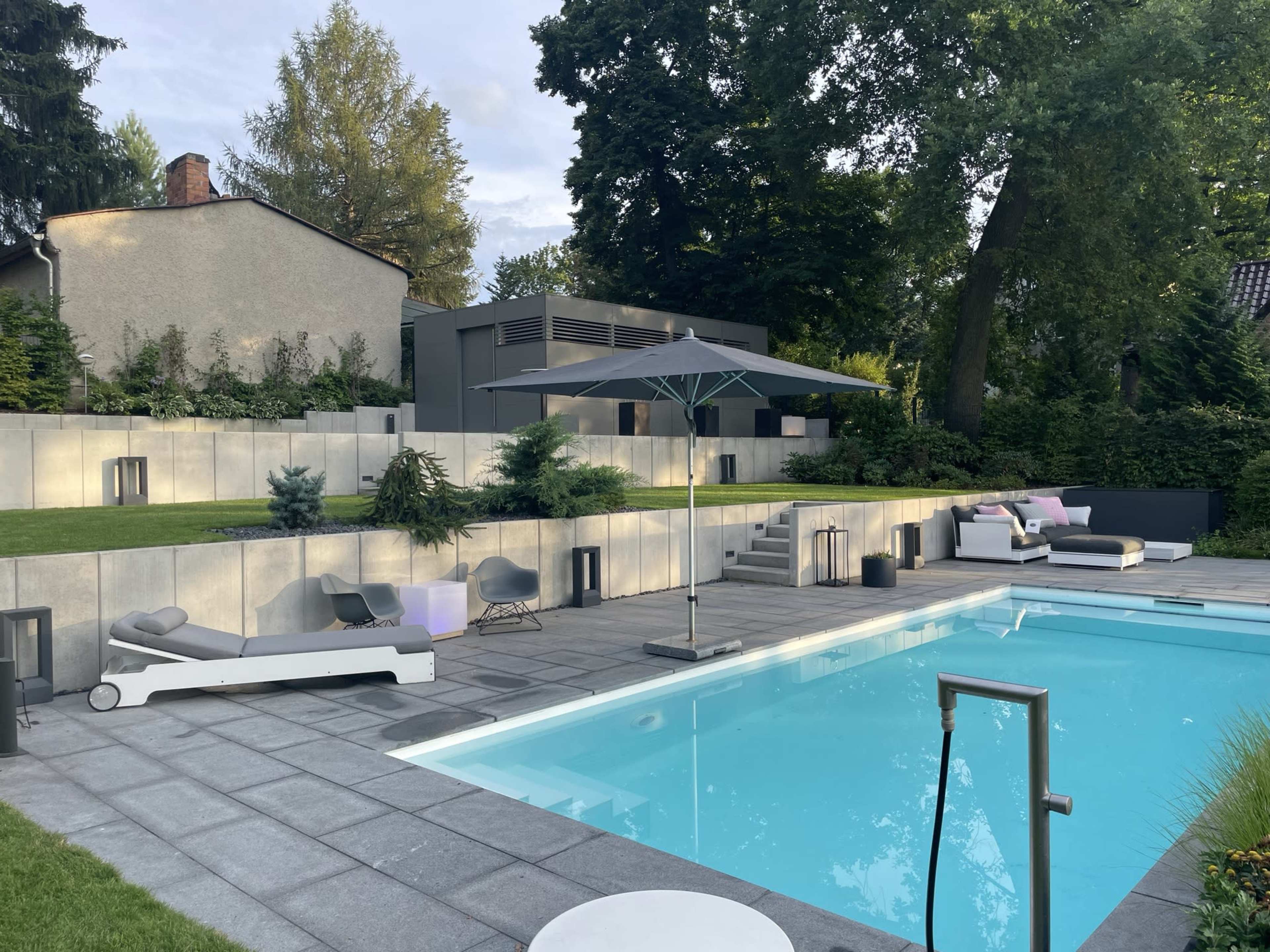 A modern backyard with a swimming pool, lounge chairs, an umbrella, and landscaped greenery surrounding a contemporary house.