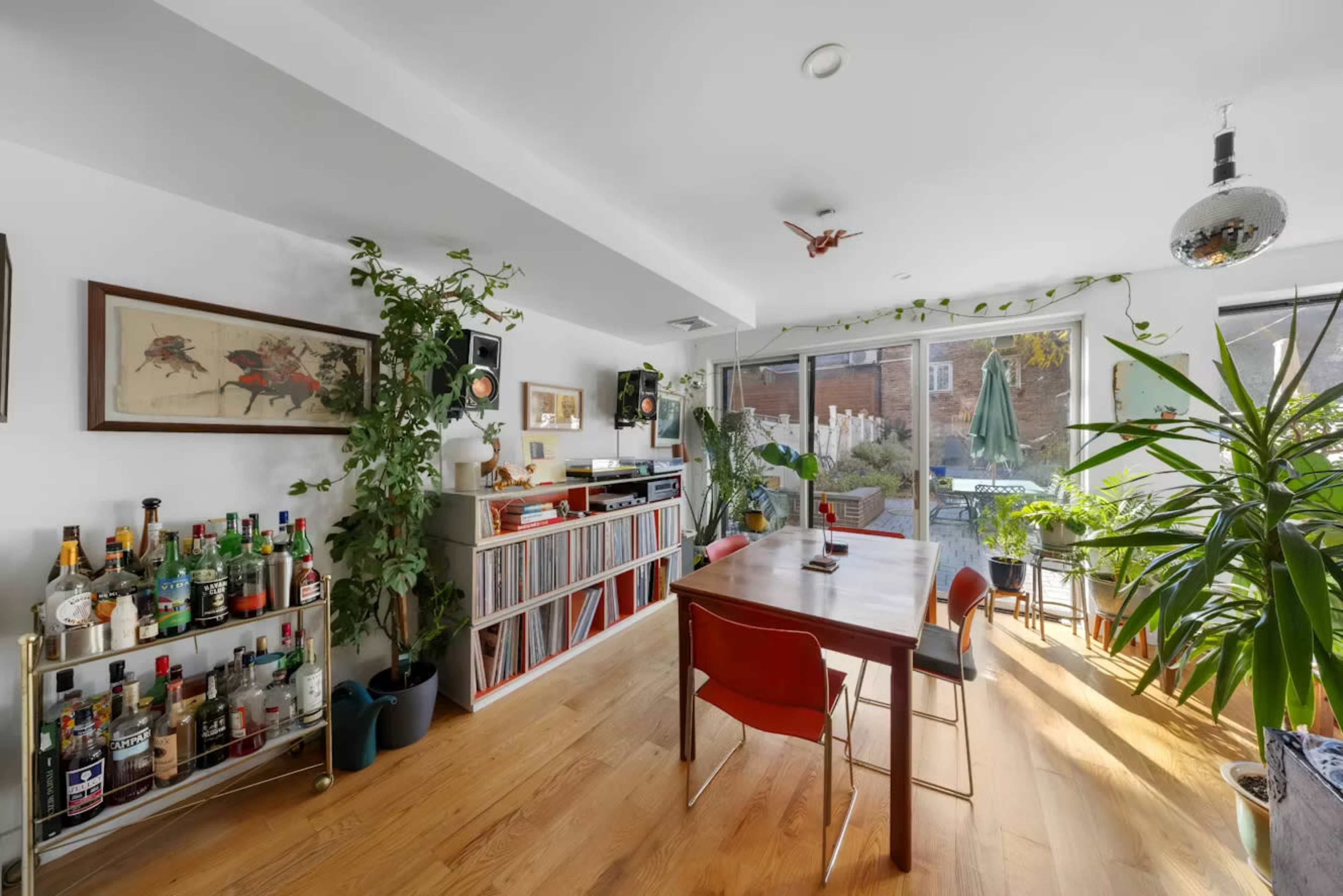The image shows a bright living space with a wooden dining table, plants, a bar cart filled with bottles, and large windows overlooking a garden.