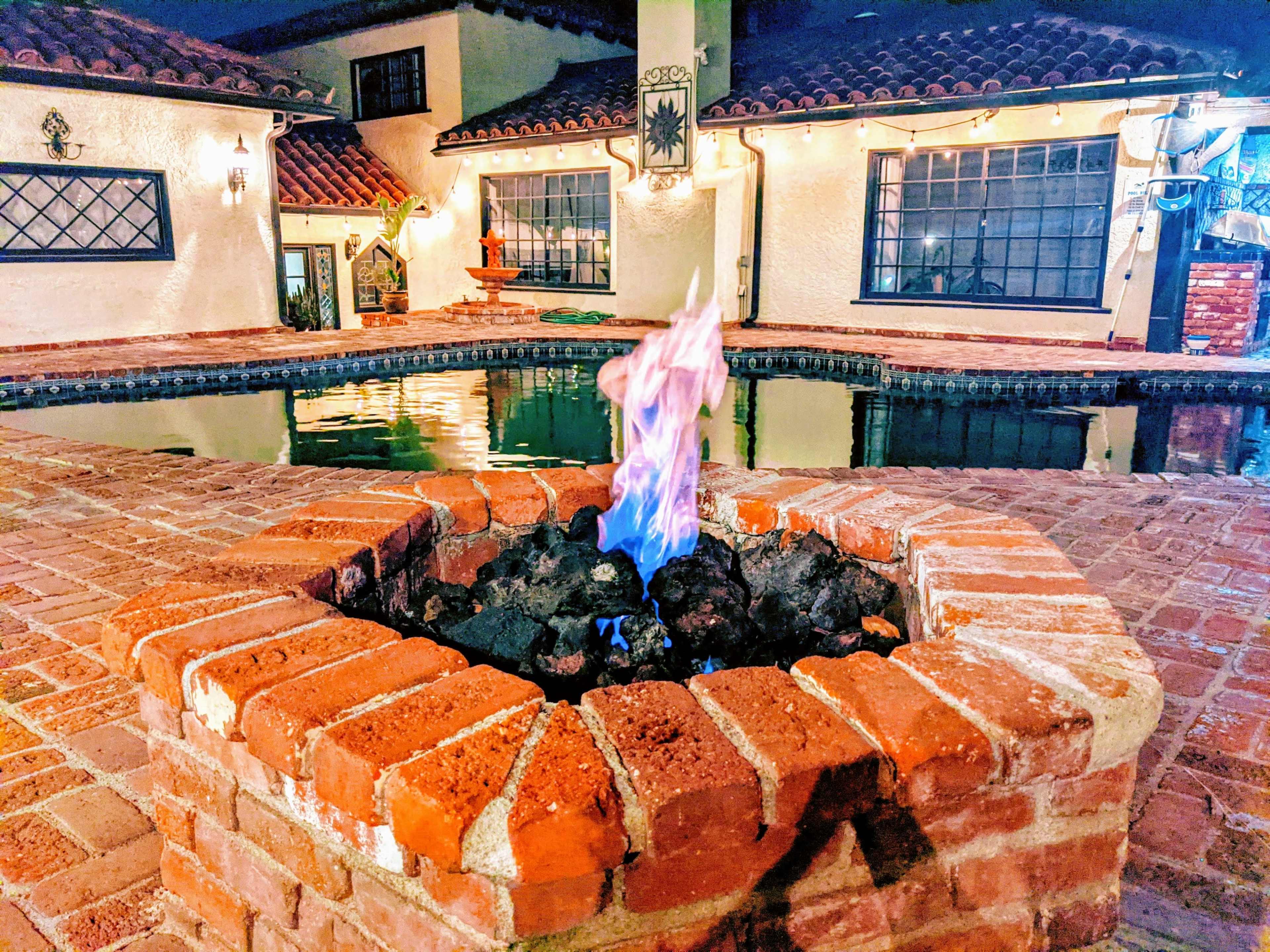 A brick fire pit with blue and orange flames is surrounded by a rectangular pool and a patio area with lights strung overhead.