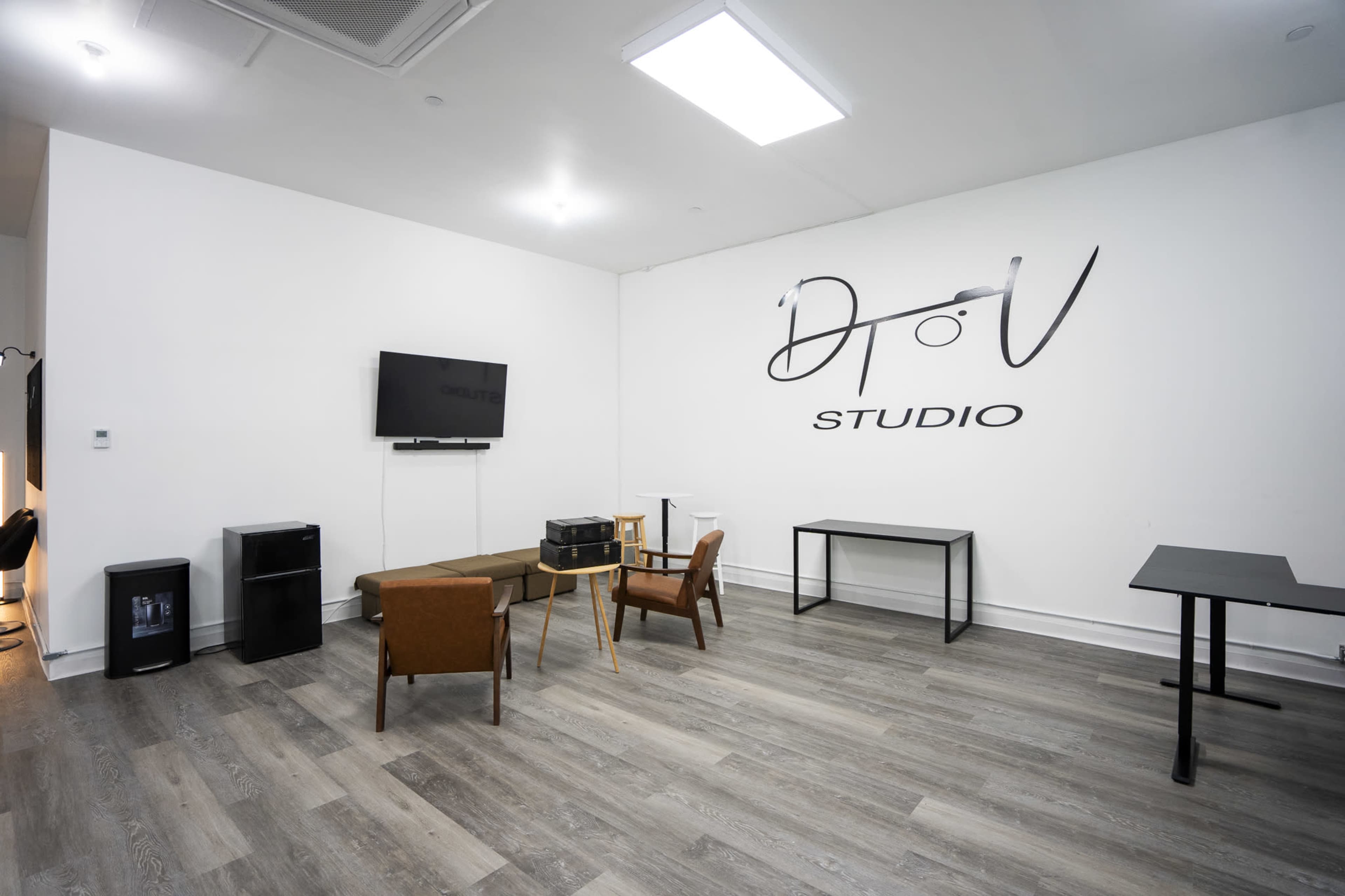 The image shows a minimalist studio space featuring a wall-mounted TV, a brown couch, two chairs, a small table, and a refrigerator against a white wall with "DToV STUDIO" written on it.