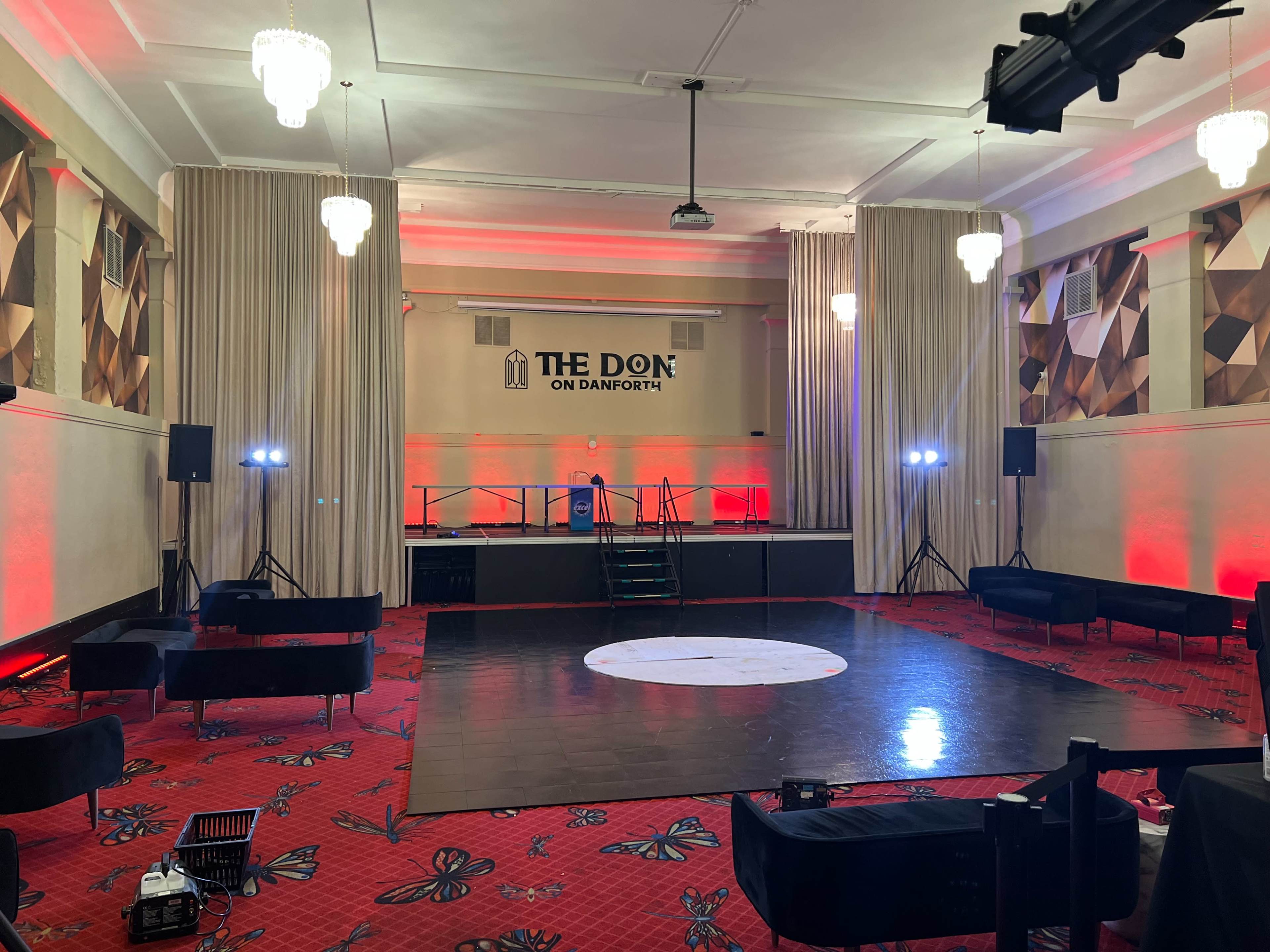 A dimly lit event space with a central stage area surrounded by chairs and audio-visual equipment.