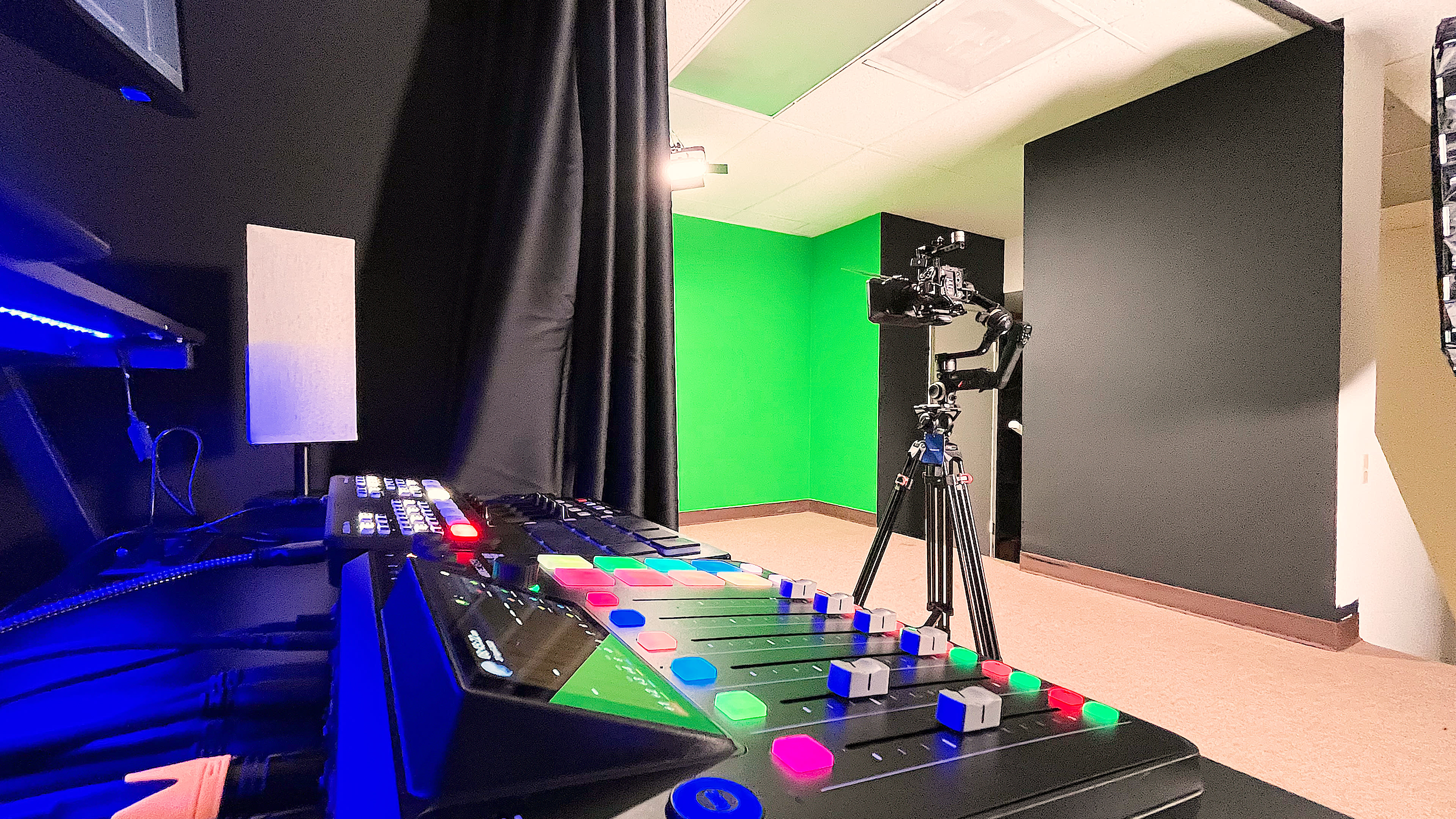 The image shows a studio setup featuring a black control panel with colorful buttons in the foreground and a camera on a tripod facing a green screen backdrop.