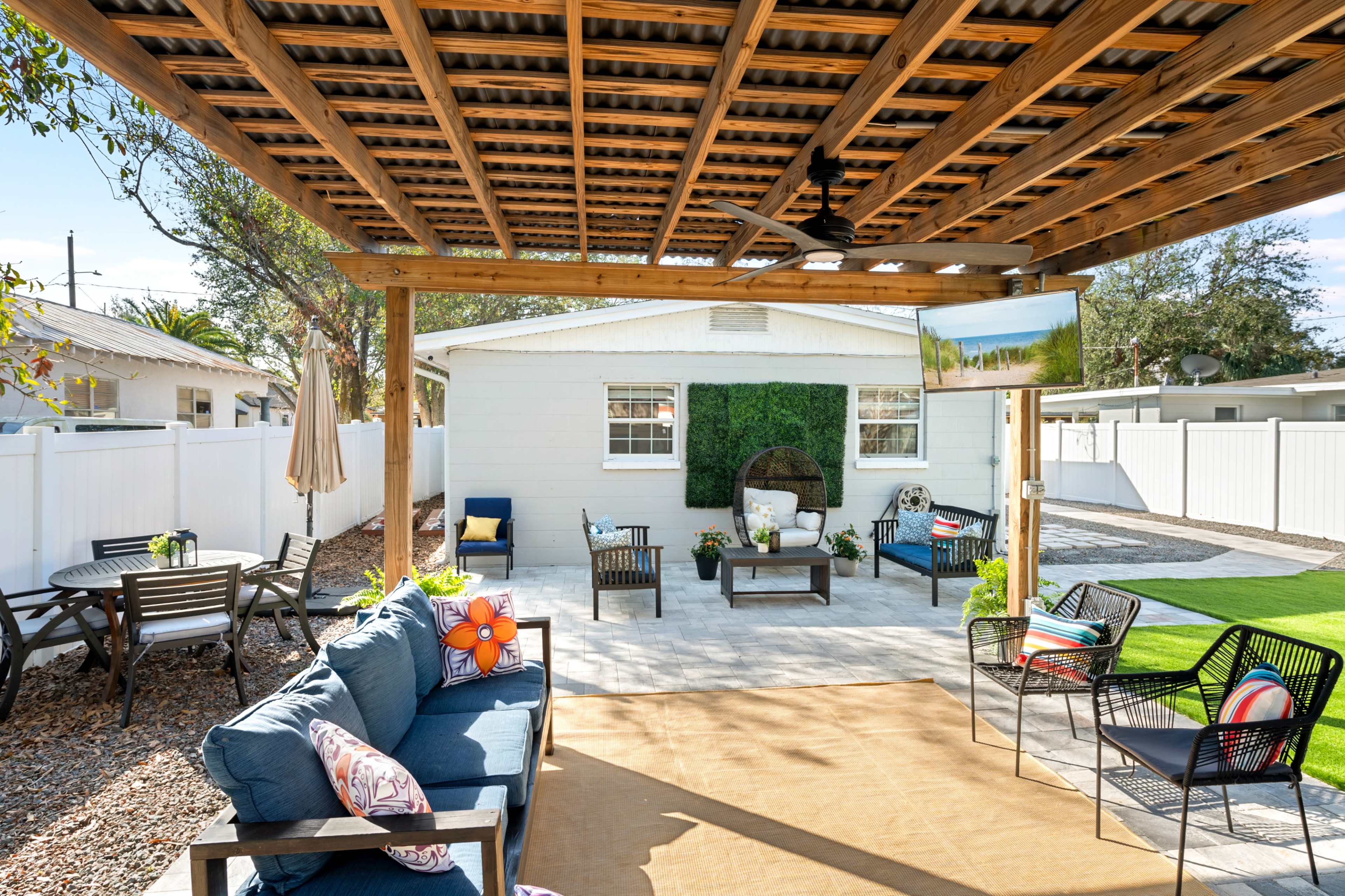 The image shows a patio area with a covered seating space, featuring a sofa, chairs, and a television, alongside a lawn and a white-walled house in the background.