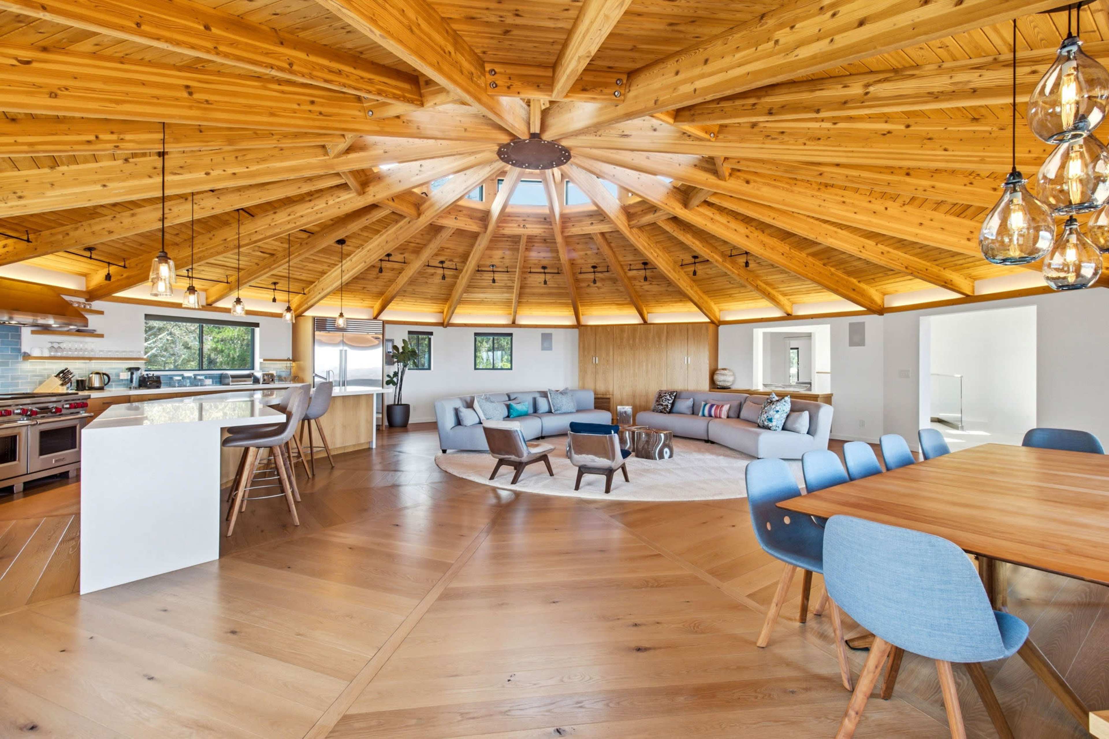 The image features a spacious, circular living area with a wooden ceiling and modern furnishings, including a large sofa, dining table, and an open kitchen area.