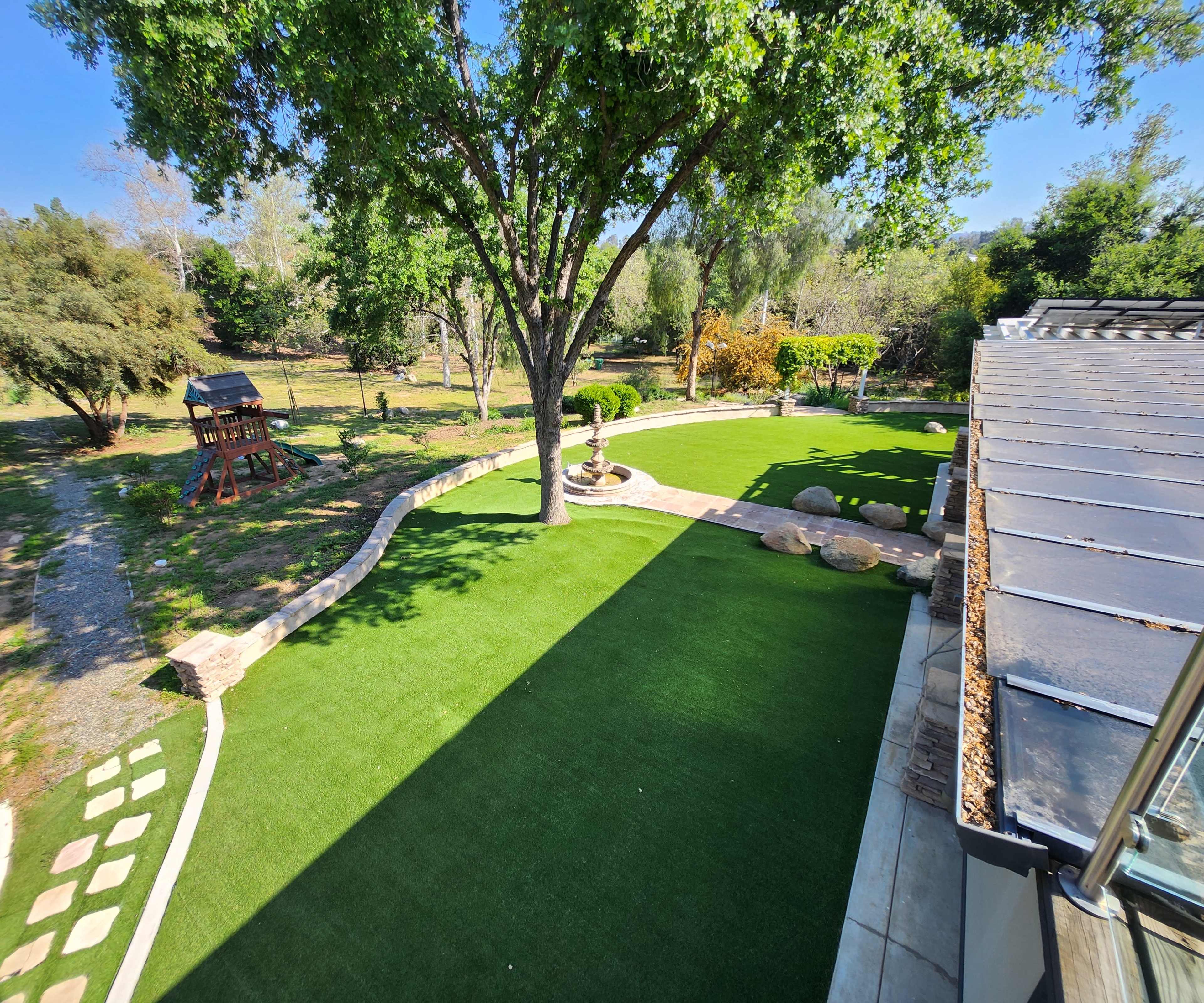 The image shows a landscaped yard with artificial grass, a stone pathway, and a large tree, surrounded by various shrubs and a wooden play structure in the background.