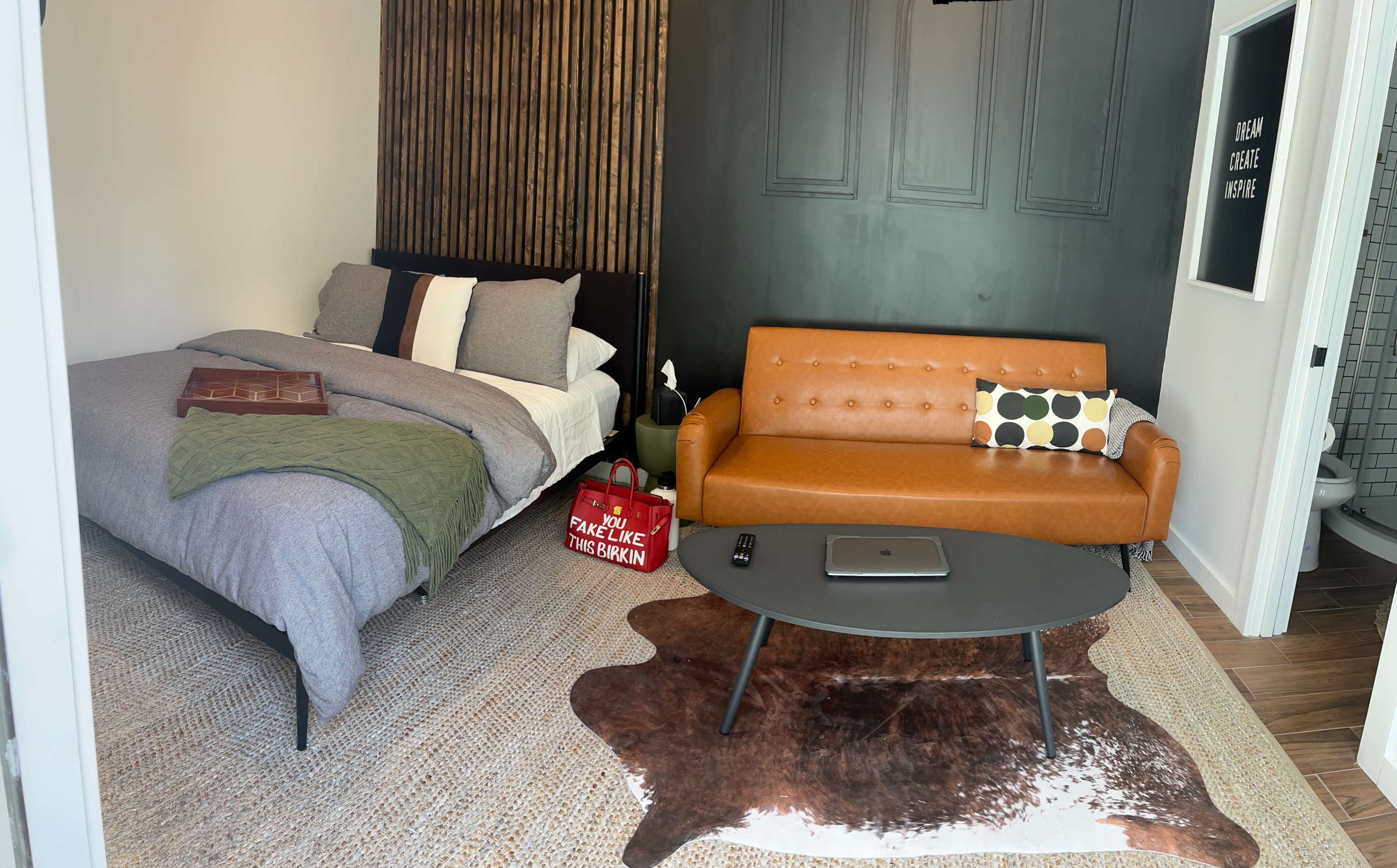 A small room with a bed on one side, a brown leather sofa, a coffee table, and a textured rug on the floor.