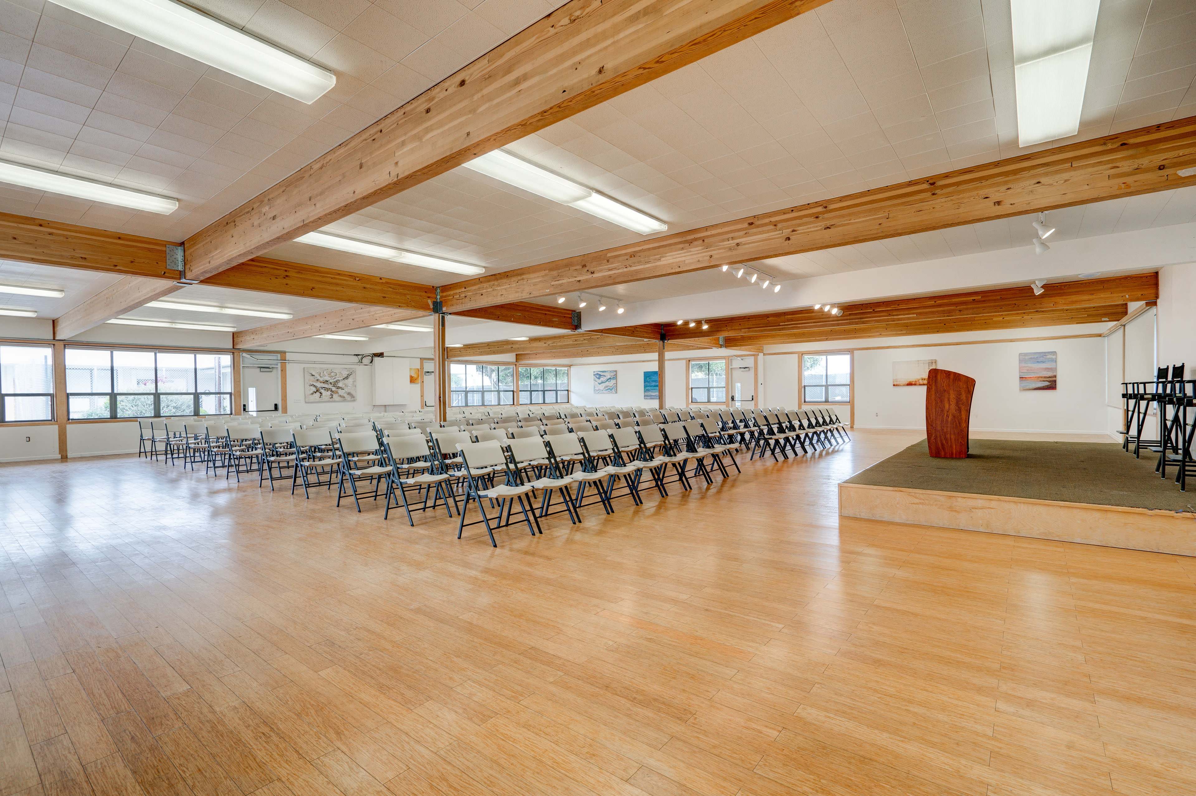 A spacious interior room features rows of chairs facing a podium on a raised platform with large windows allowing natural light.