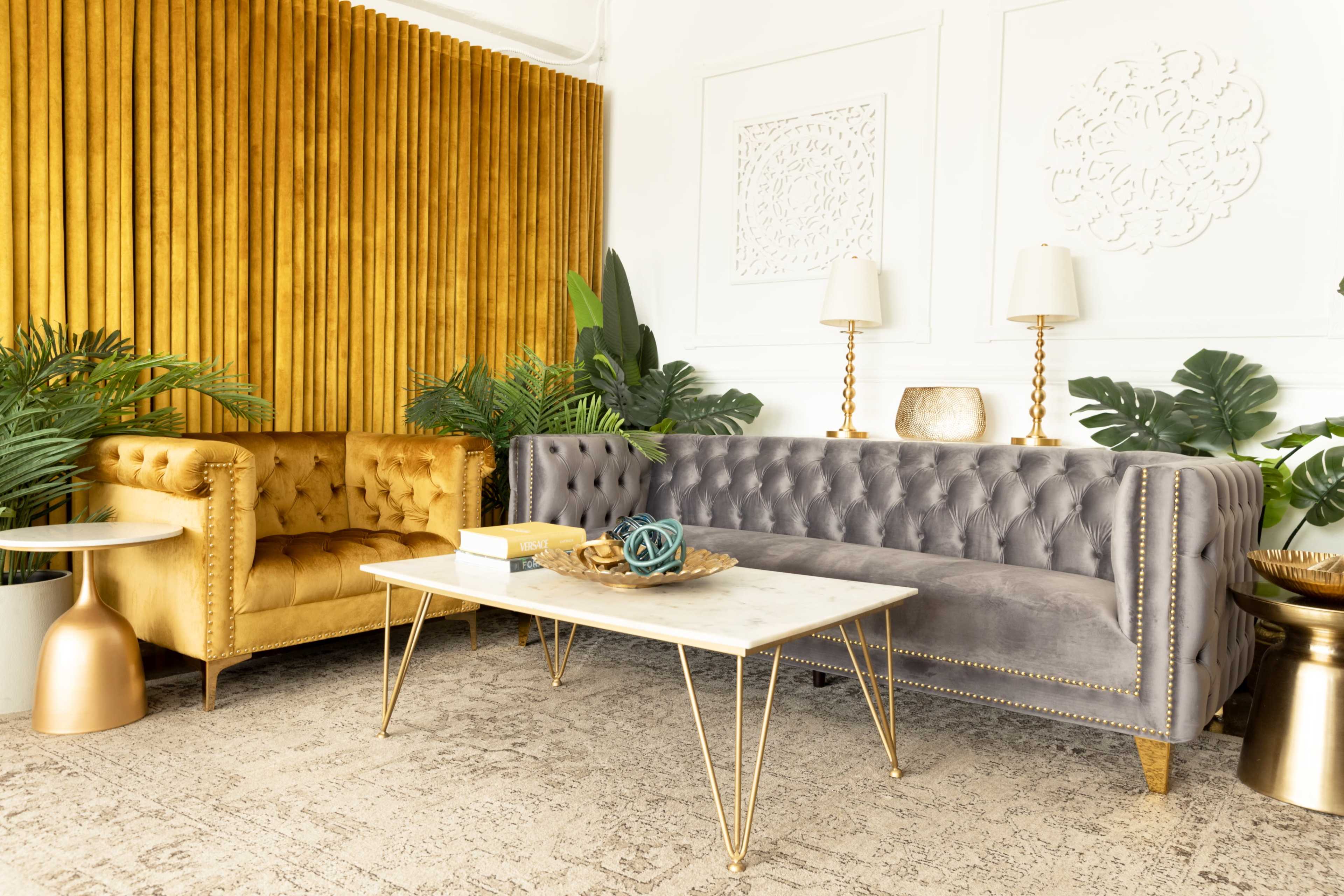 The image shows a stylish seating area with a gray tufted sofa, a golden armchair, a marble coffee table, and lush greenery against a textured wall.