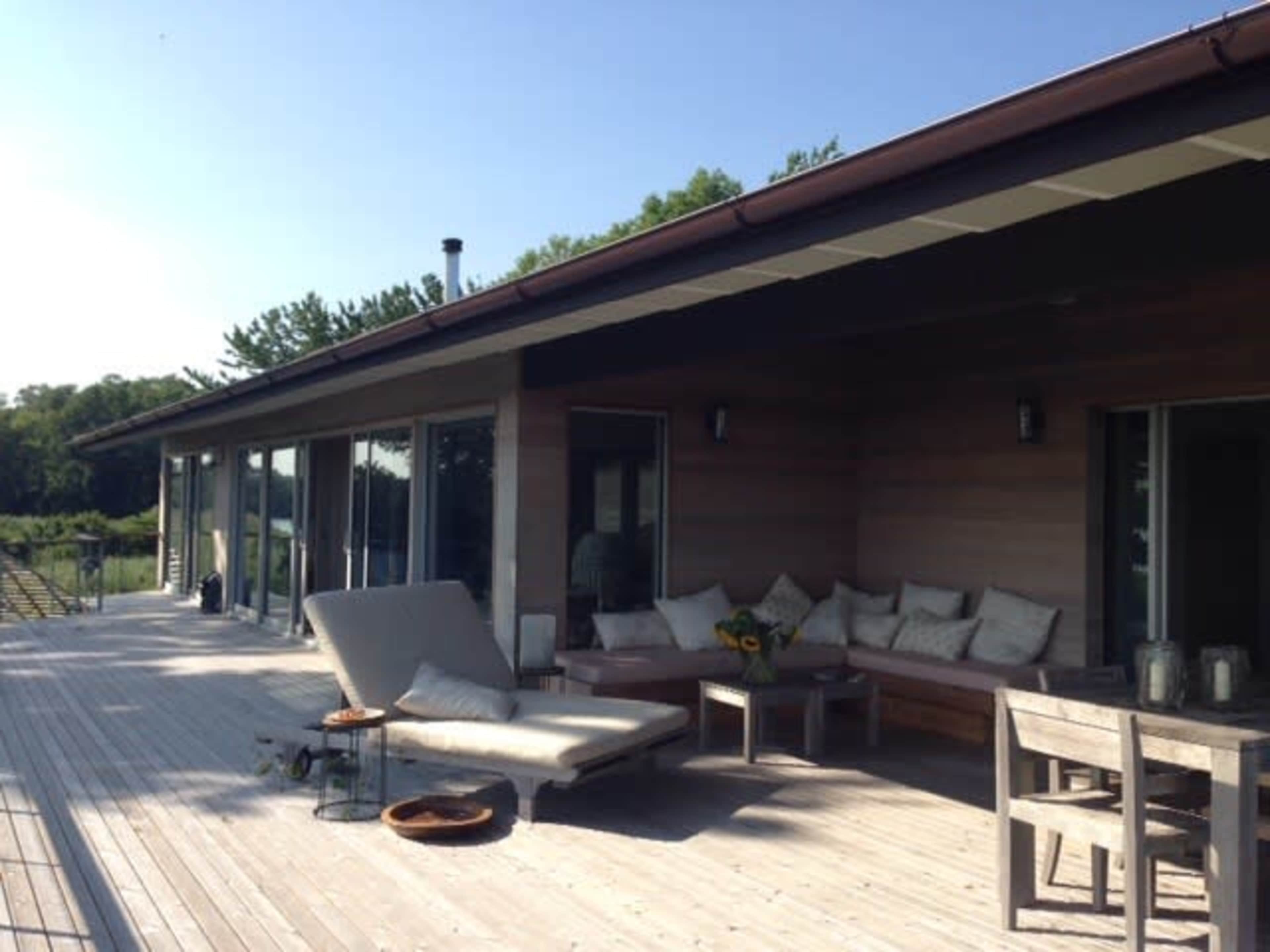 A modern wooden deck features a lounge chair, a sectional sofa, and a dining table, overlooking a green landscape.