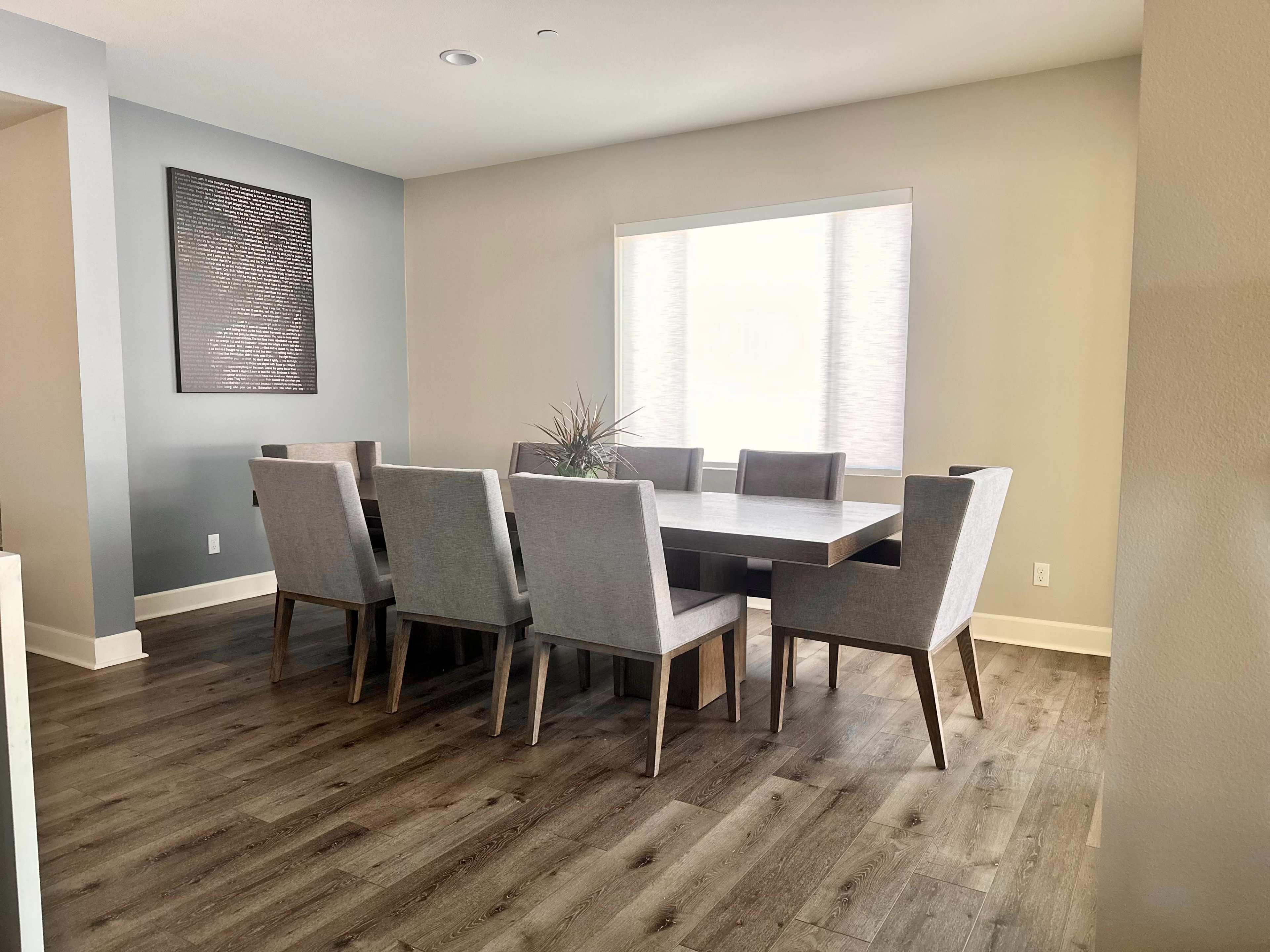 A modern dining room features a large rectangular table surrounded by eight grey upholstered chairs, with a bright window providing natural light.