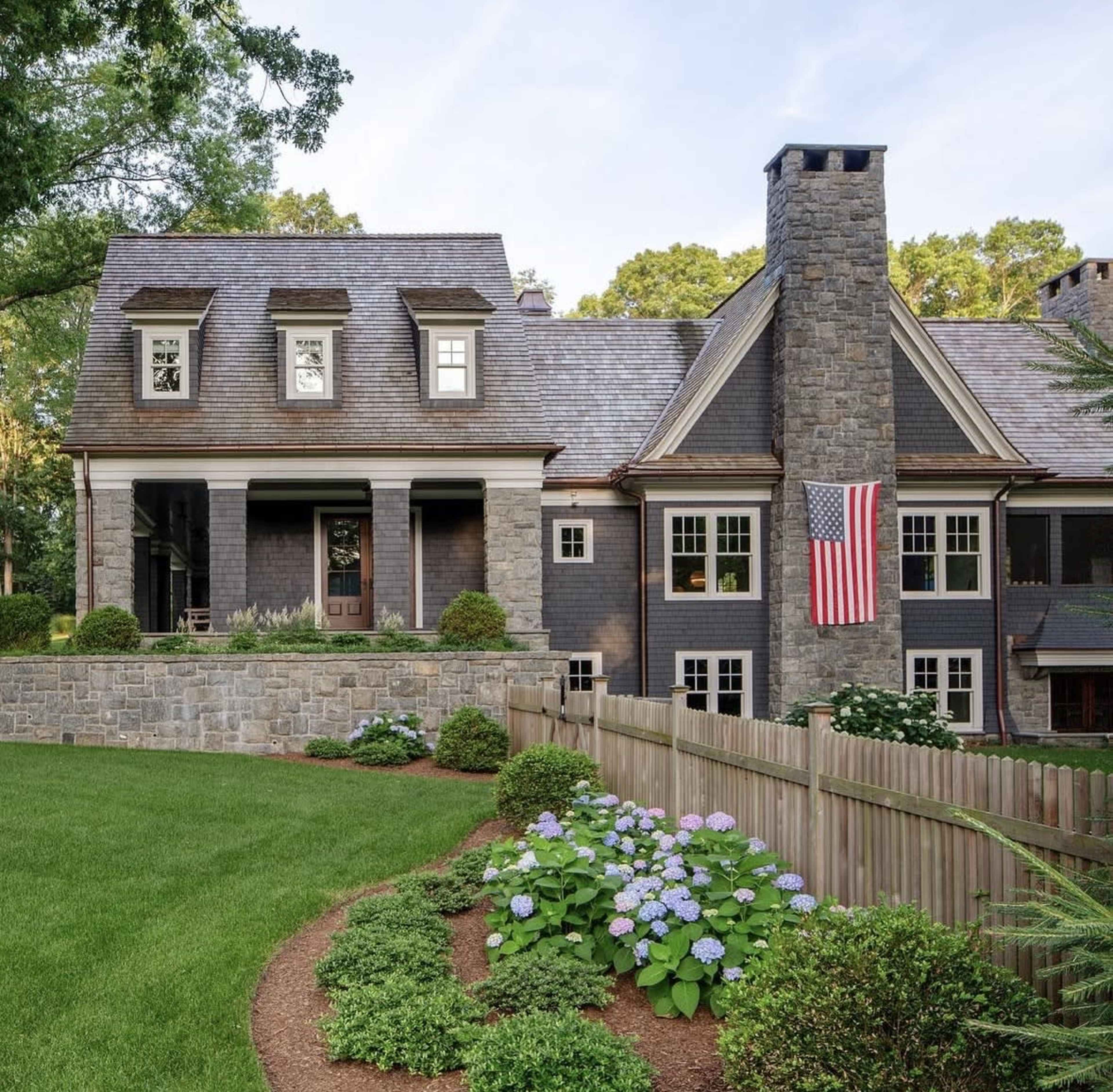 The image shows a large, two-story house with a combination of stone and dark siding, featuring a chimney and an American flag hanging from the side.