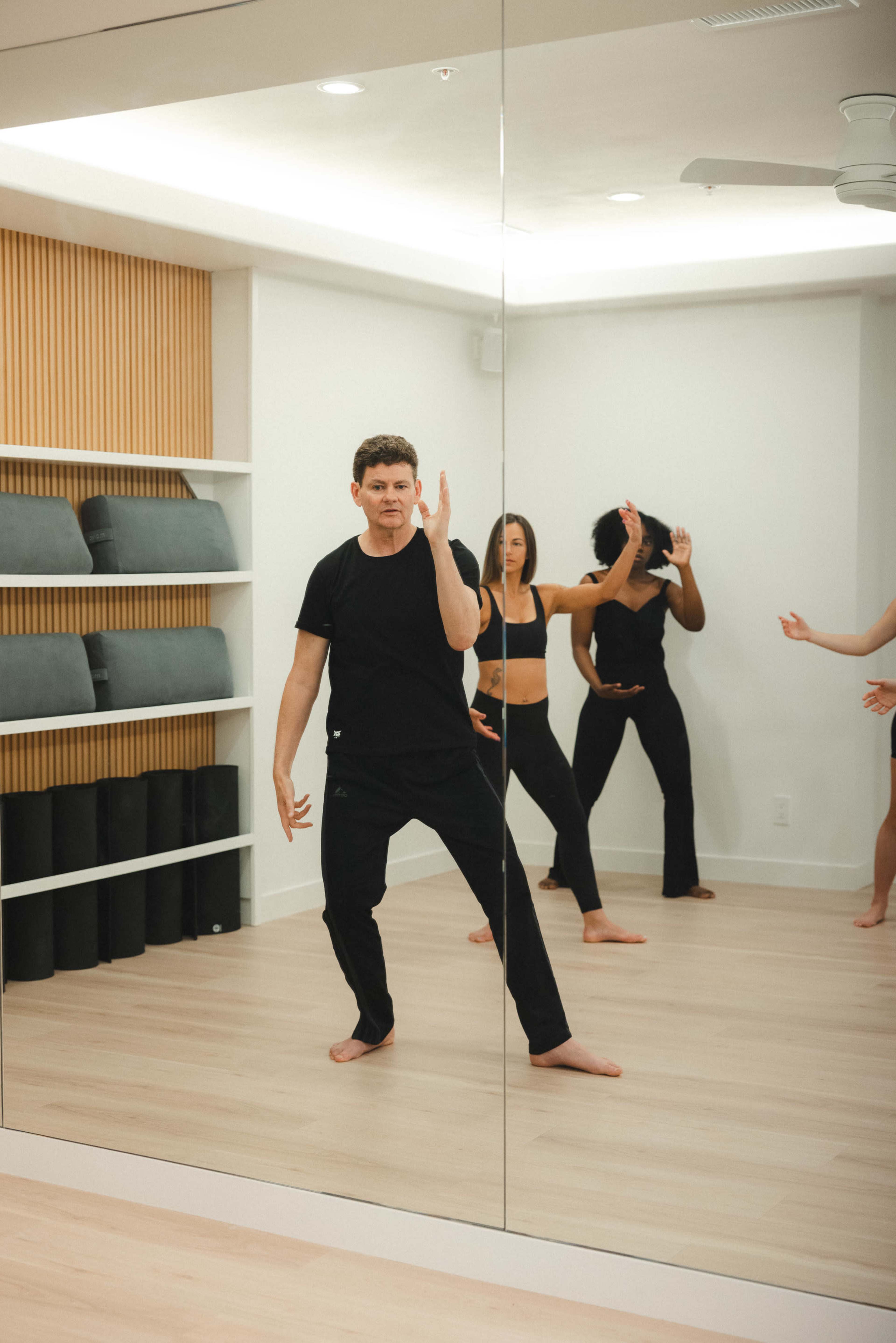 A group of individuals in black workout attire is practicing movements in a mirrored exercise studio.