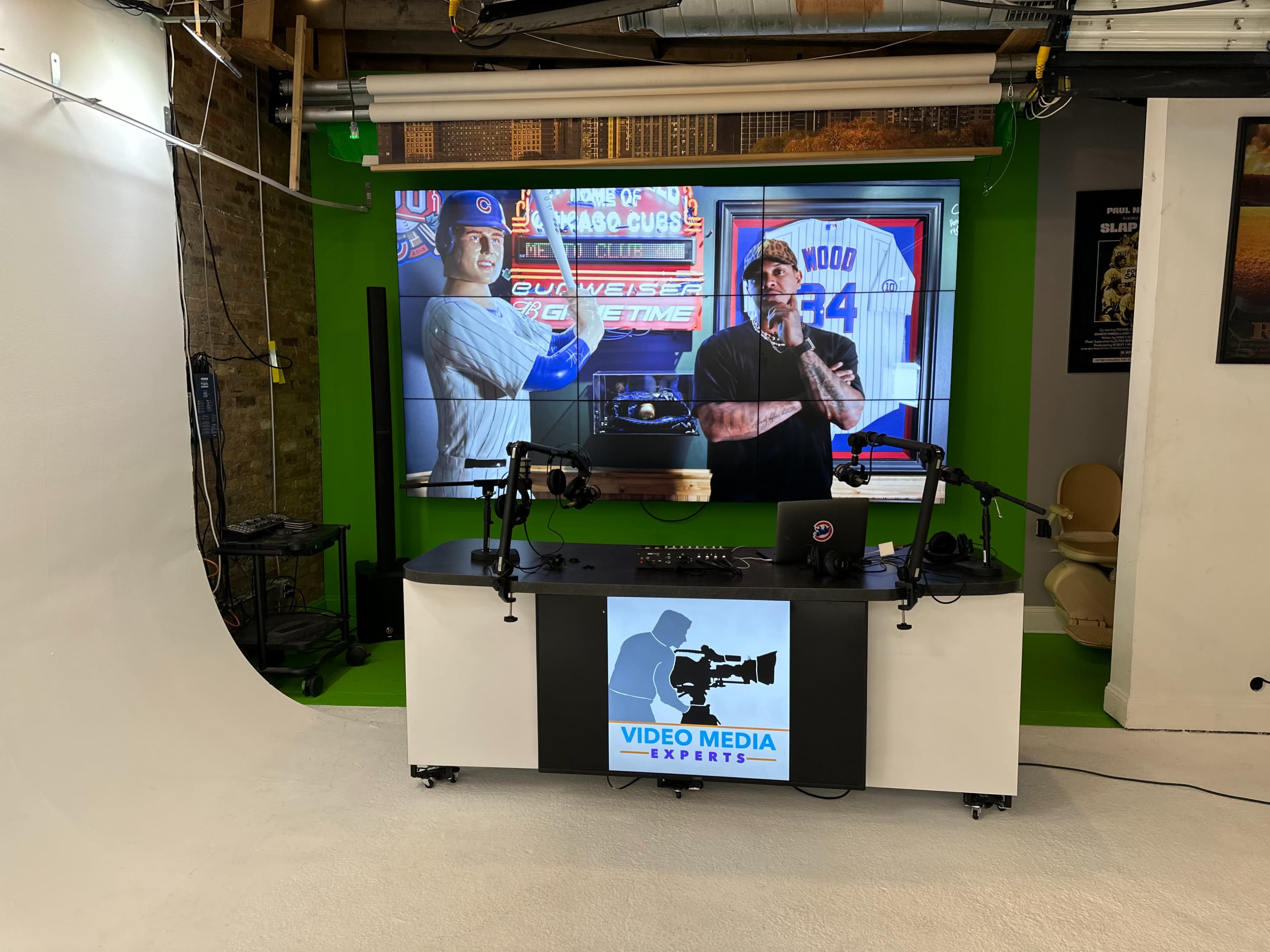 A video recording studio with a large screen displaying sports images, a central desk equipped for broadcasting, and various production equipment in the background.
