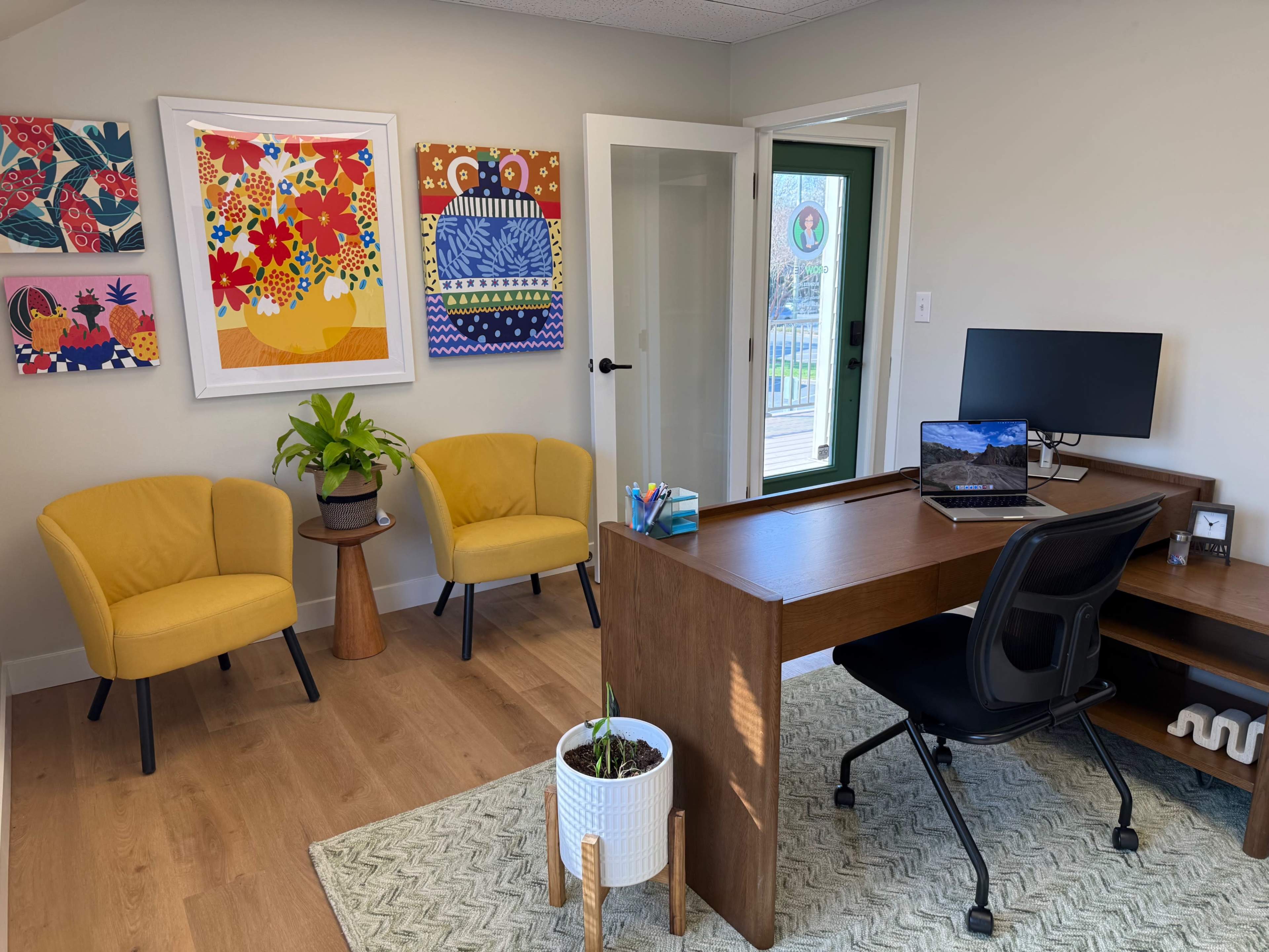 The image shows a modern office space with a wooden desk, a computer, two yellow chairs, a potted plant, and colorful artwork on the walls.