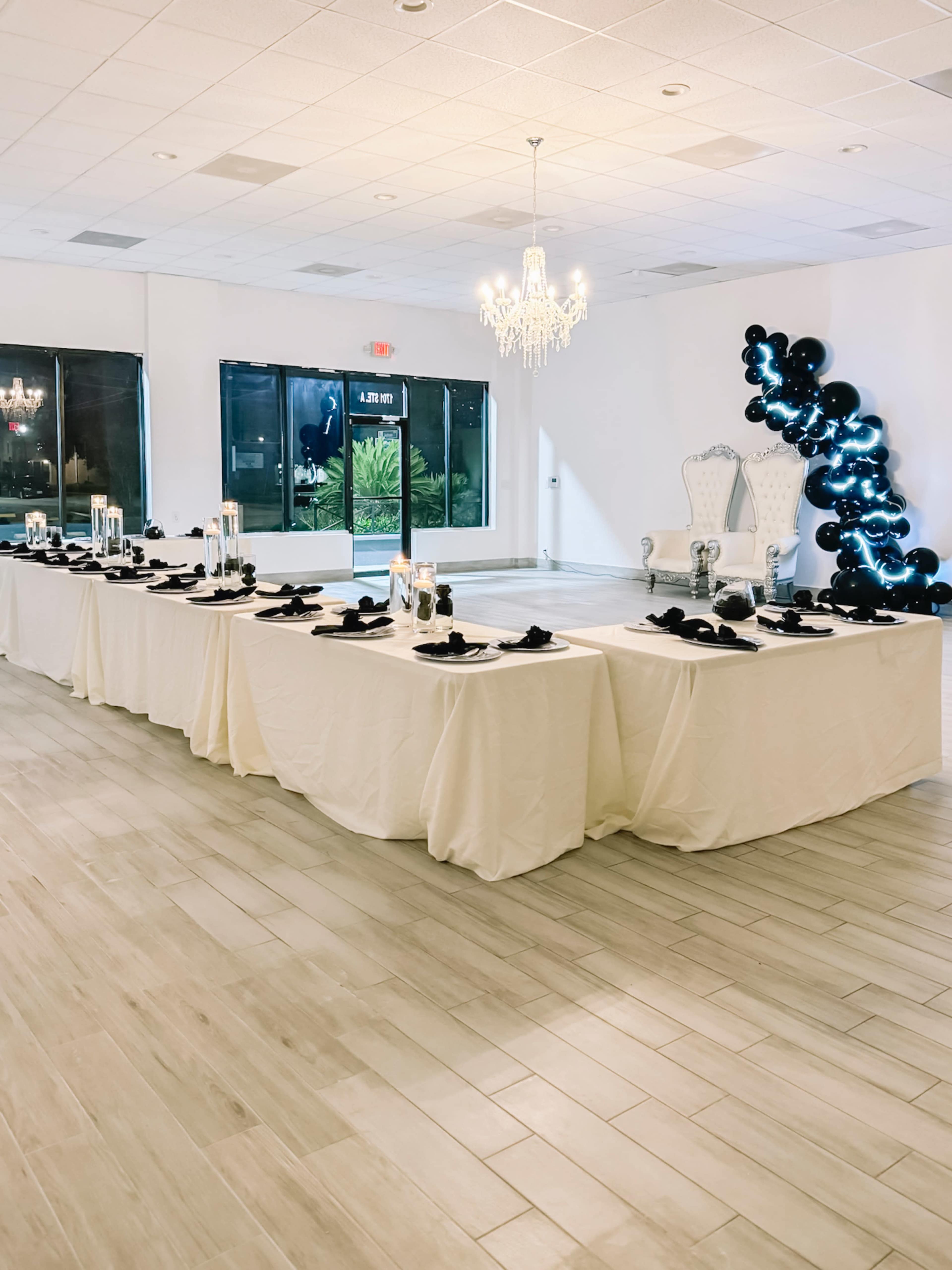 A elegantly set banquet table with white linens, black tableware, and a whimsical black balloon arch is arranged in a bright event space featuring a chandelier.